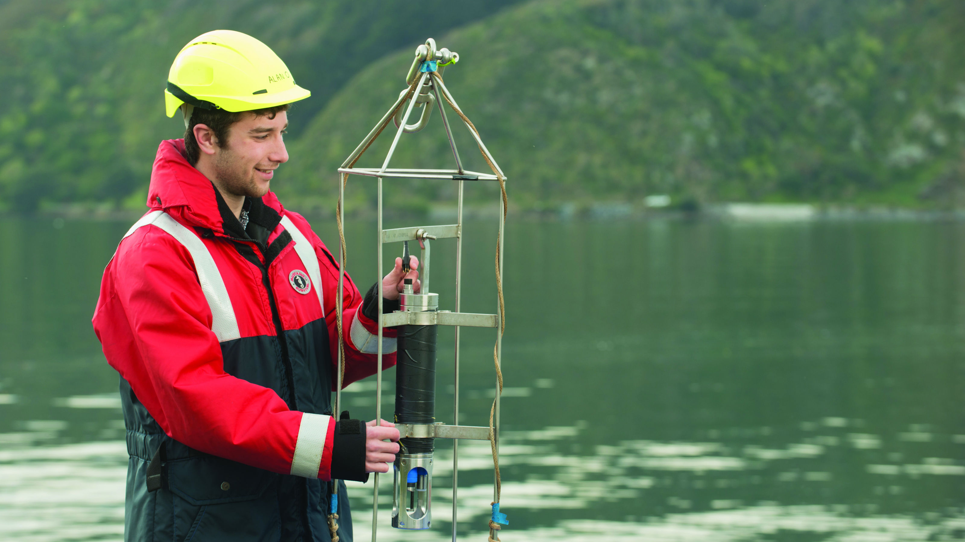 Benjamin stands in a lake with a high viz vest and a high viz helmet. He holds a scientific instrument. Image copyright Victoria University of Wellington.
