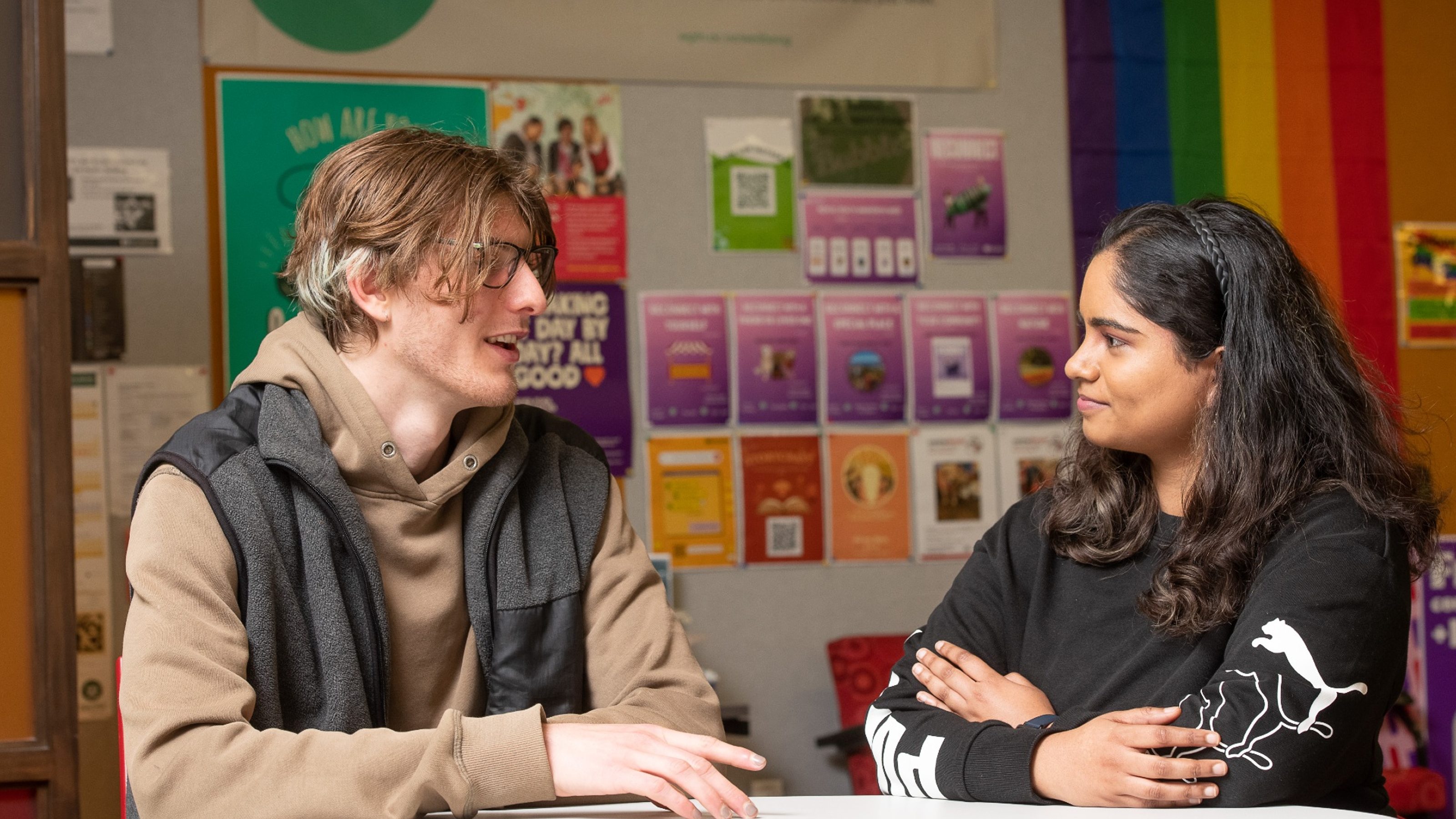 Two young people talking in the Bubble student space with posters and rainbow background.