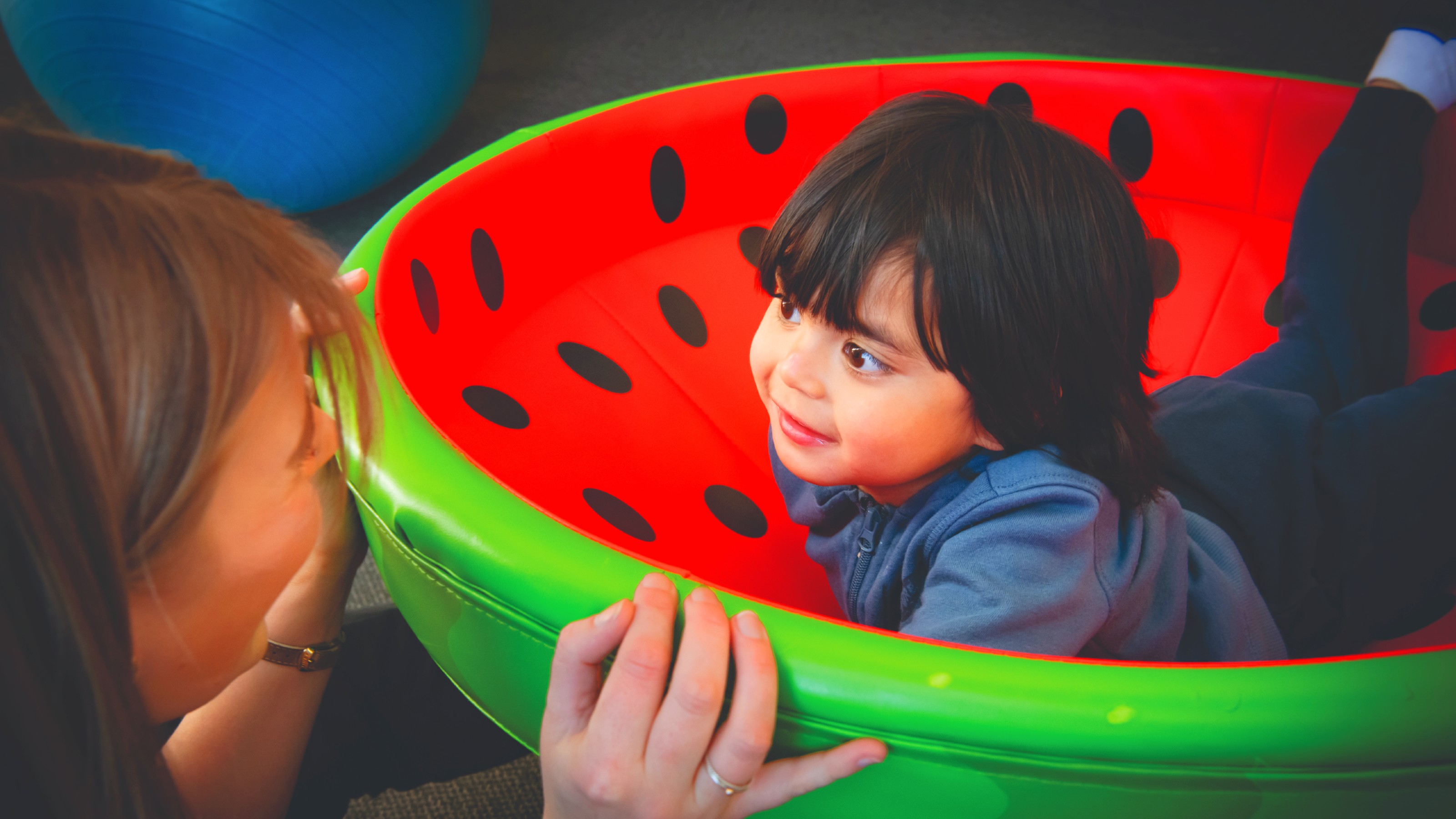 Small child lies tummy down in a red and green bowl looking at a woman