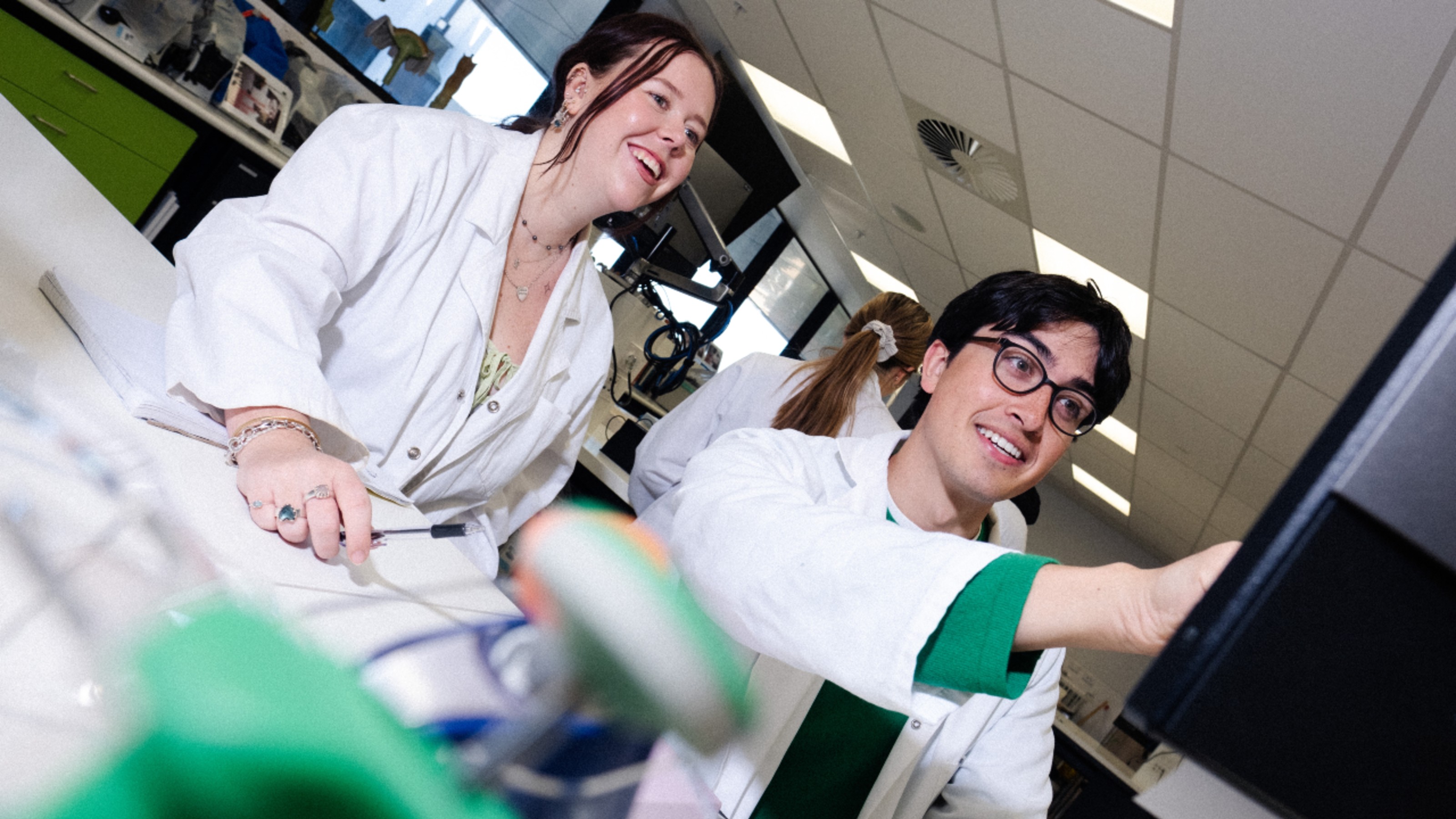 Two students in lab coats use a monitor to study Human Genetics.