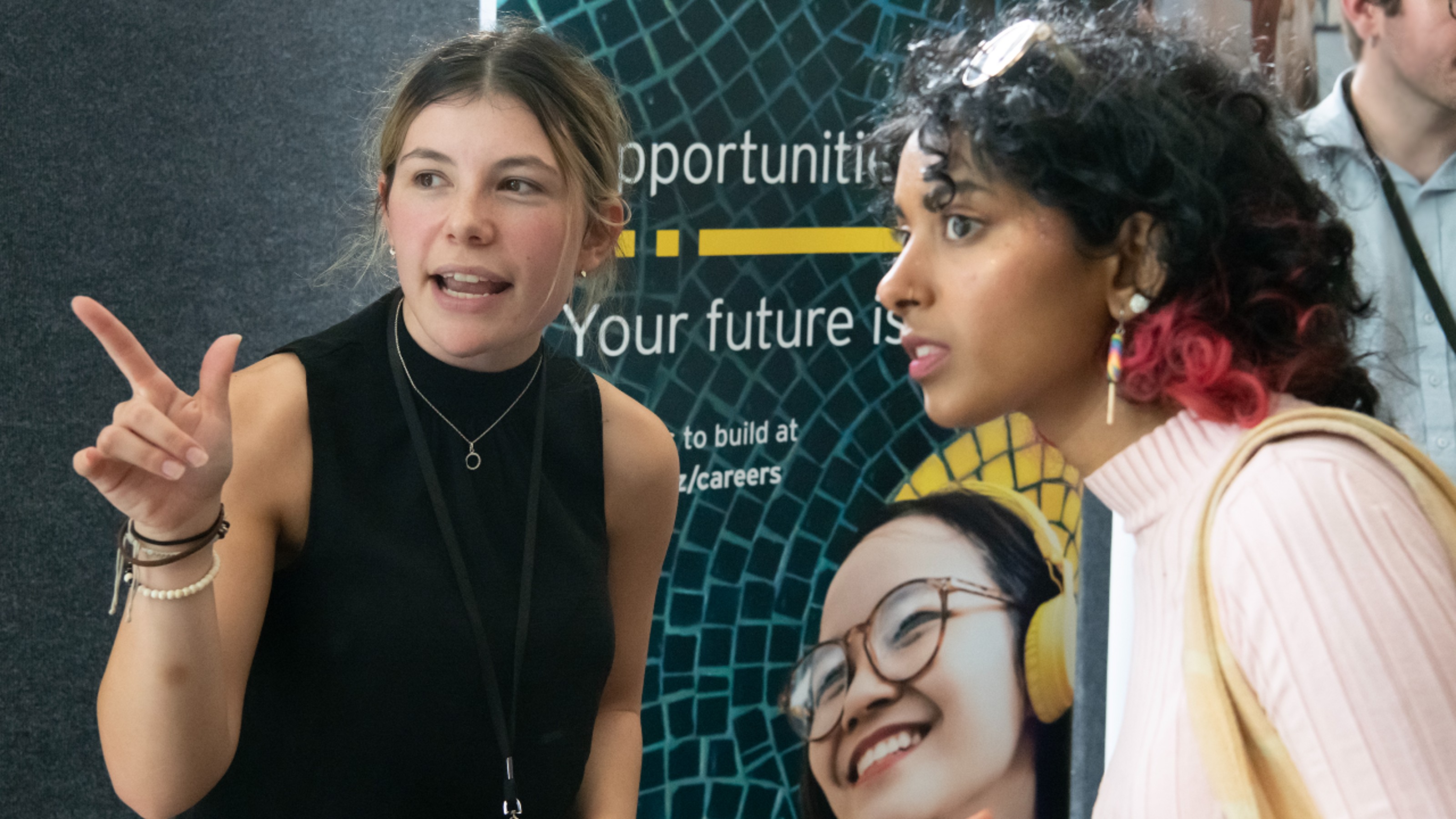 A female commerce student with pink jumper talks to a female company representative  in front of company banner at a careers expo