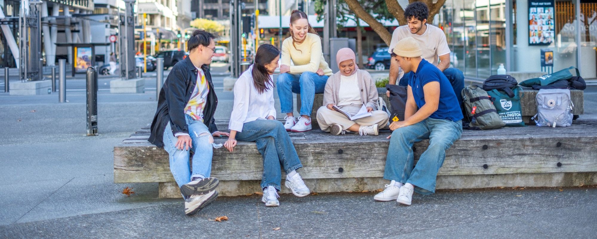 6 students sitting on a bench in the city. All students are focusing on one student who is reading from a book