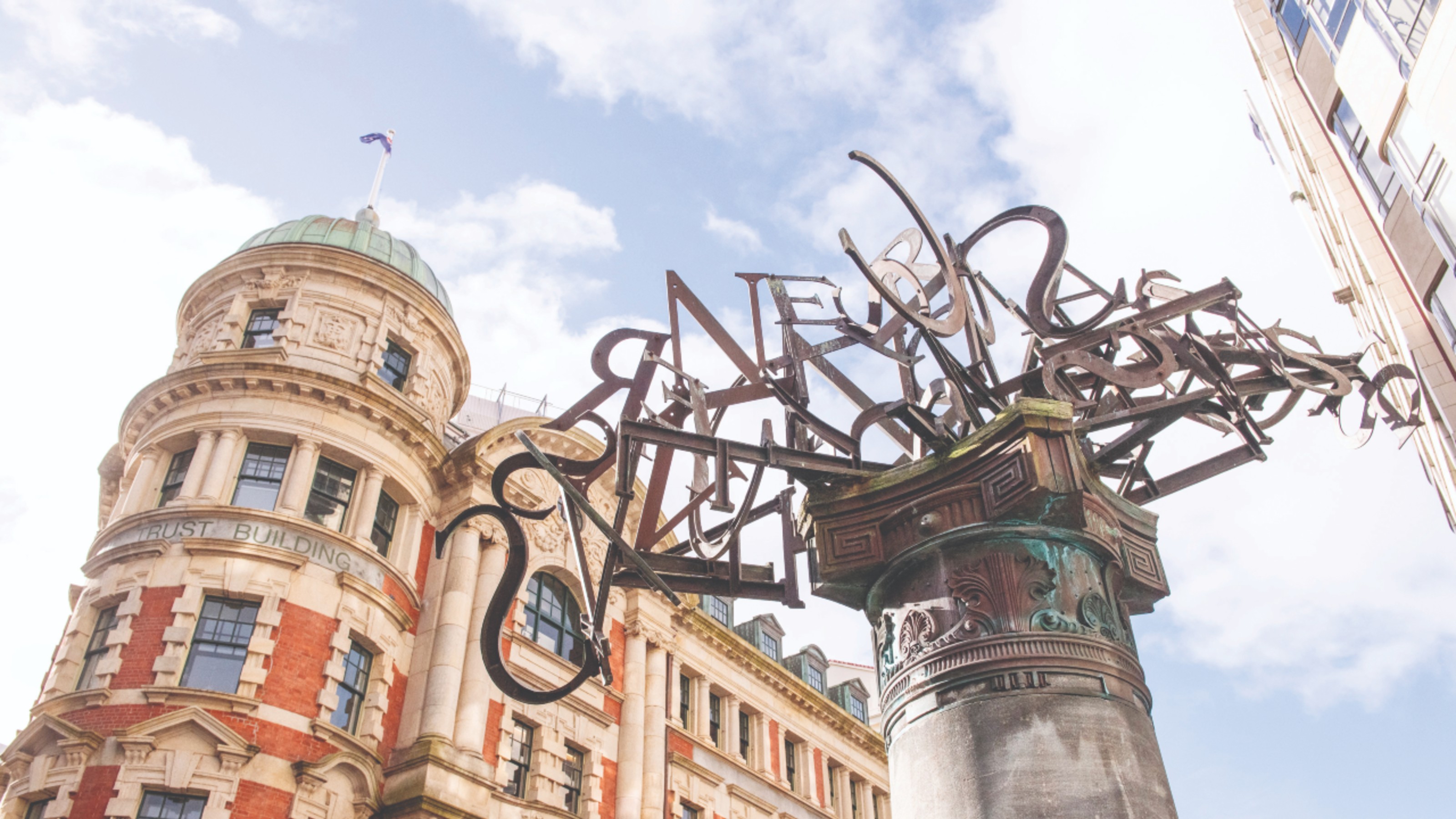 Ralph Hotere and Mary McFarlane’s ‘Rūamoko’ sculpture with the Public Trust building in the background, the sculpture features reclaimed and reused metal letters jumbled together.