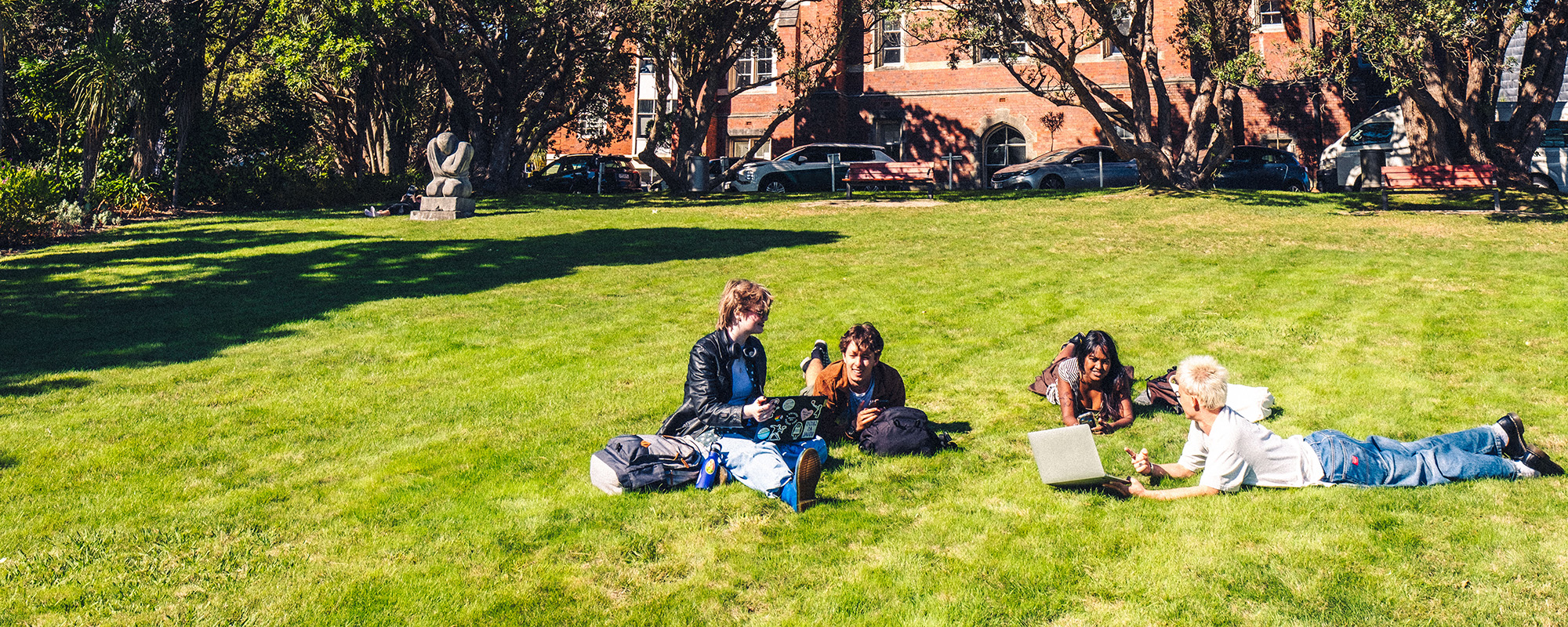 Some people sitting and lying on a lawn with laptops and backpacks talking to each other.