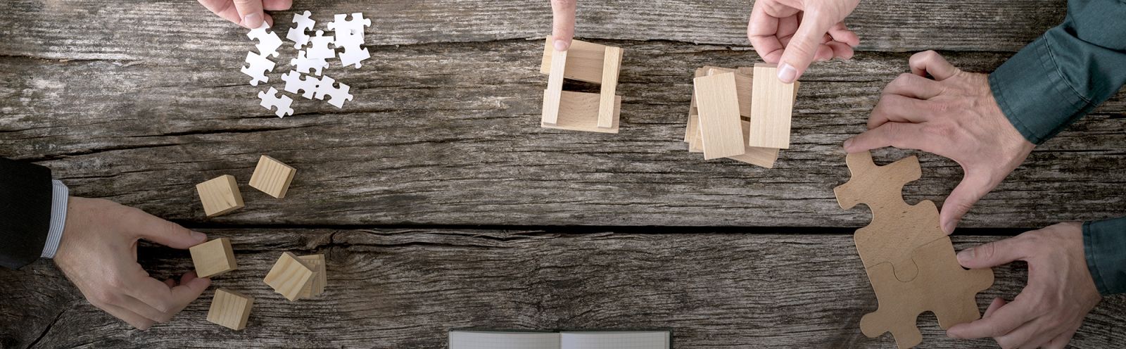 A close up of hands around a wooden table interacting with objects like cubes, puzzles, wooden prisms and puzzle pieces.