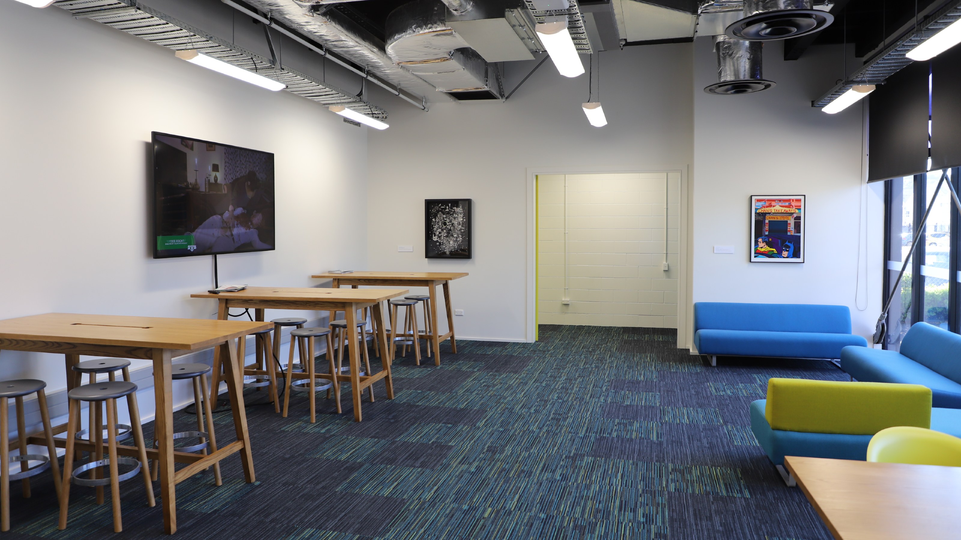 Lounge with high stools and tables to the left, and blue couch and green chairs to the right, and wide window.