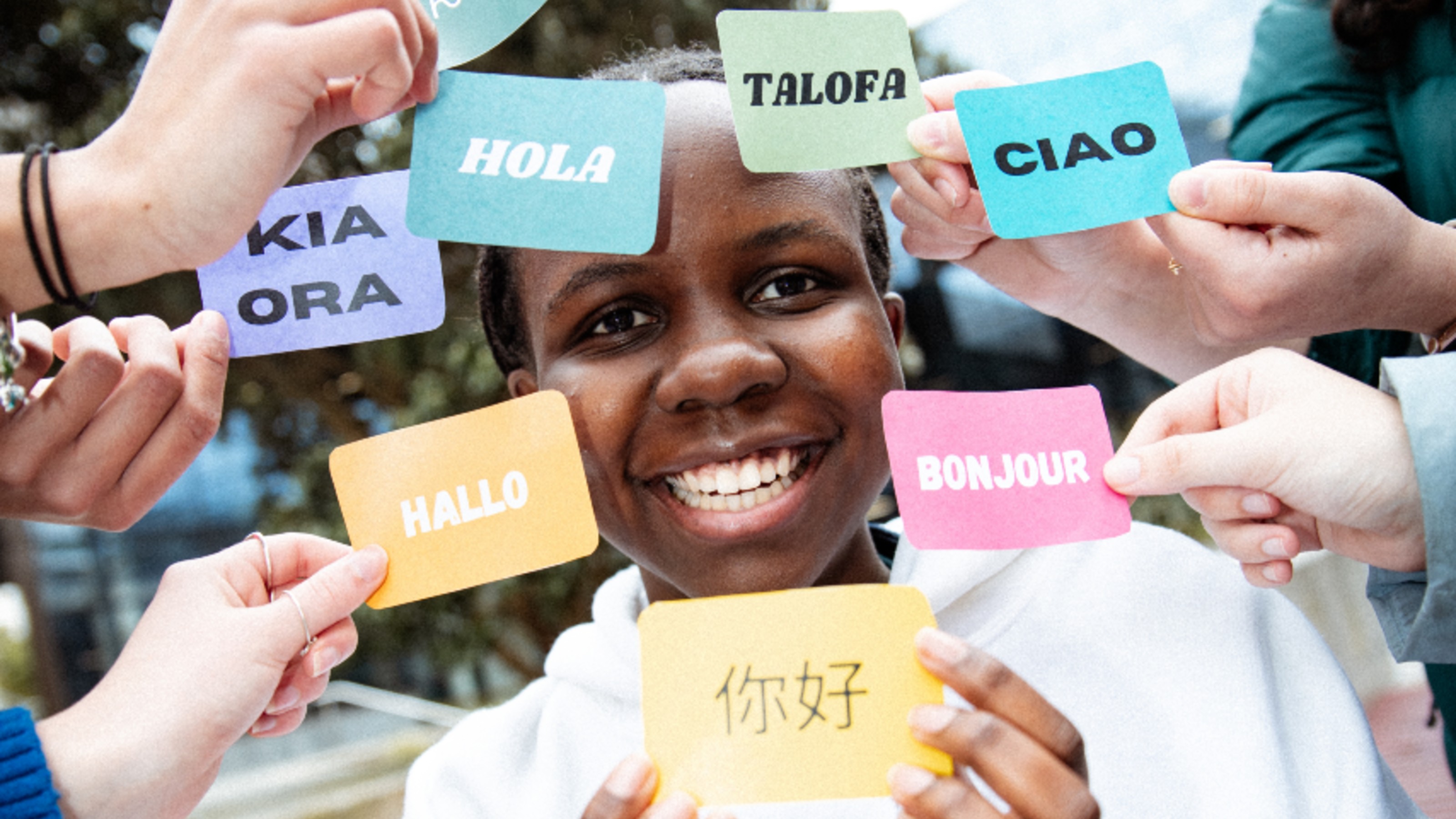 A feminine student looking at the camera with people holding cards around her face showing 'hello' in multiple languages.