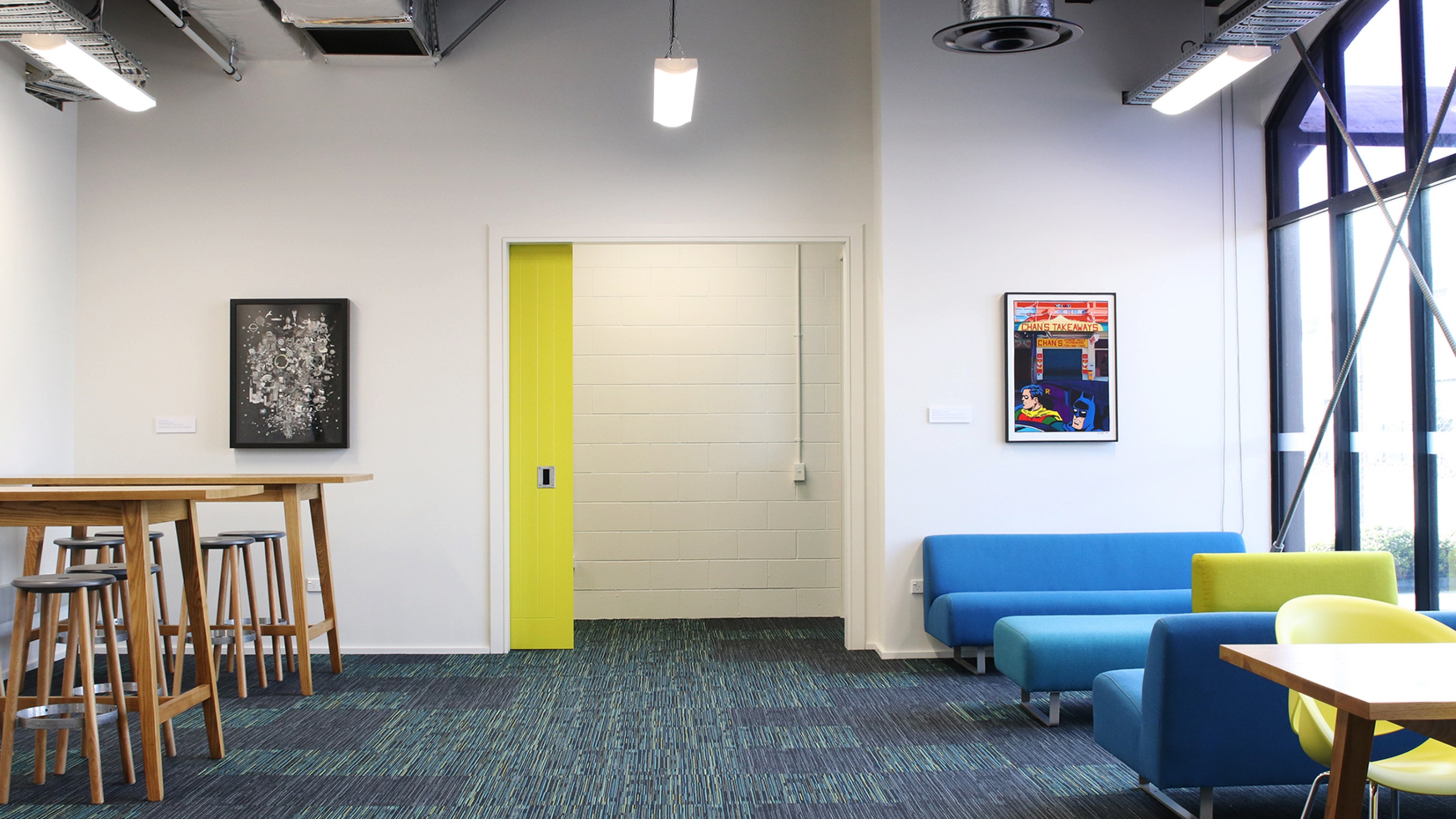 Lounge with high stools and tables to the left, and blue couch and green chairs to the right, and wide window.