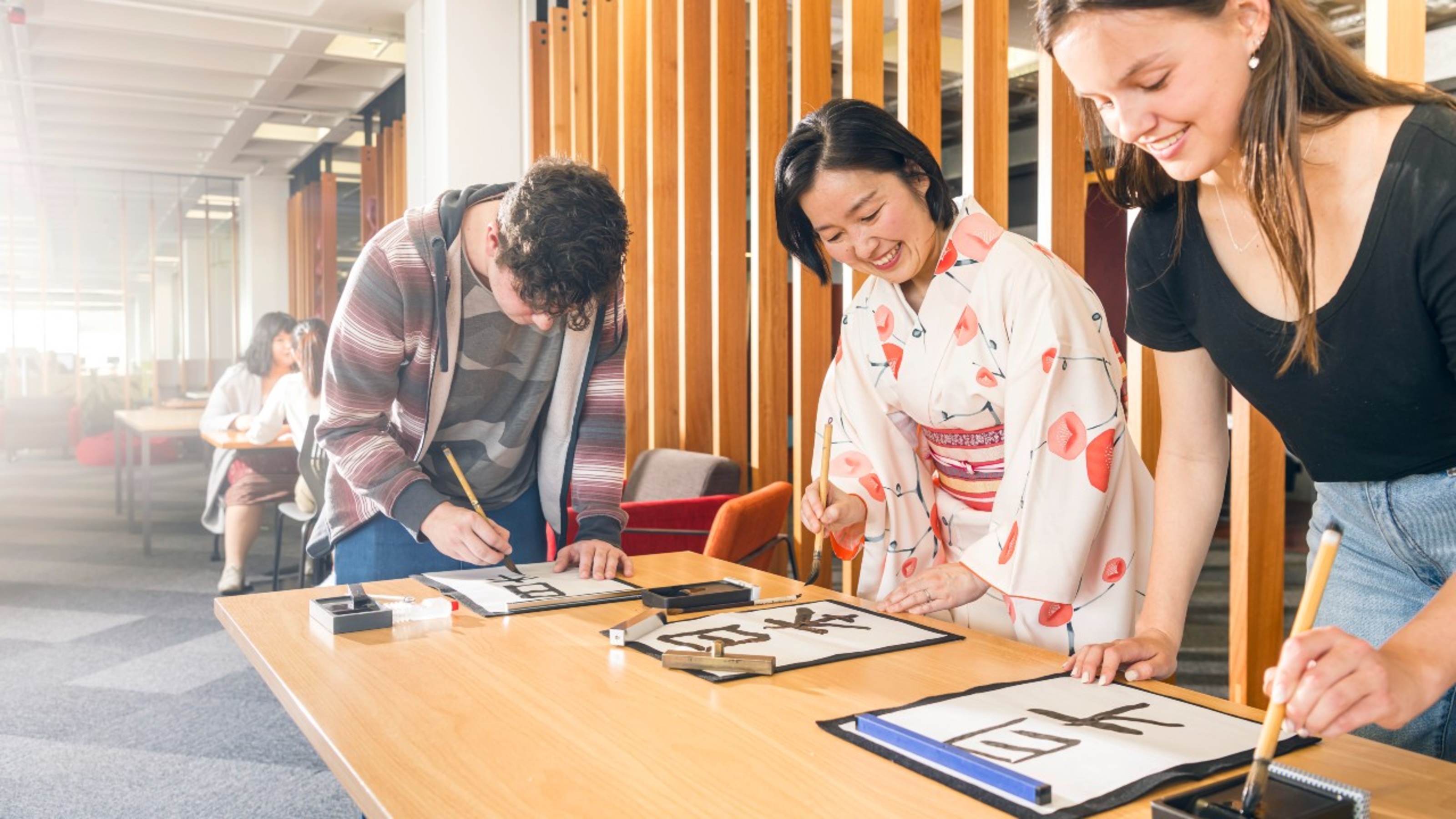 Students practicing calligraphy with a tutor who is wearing a kimono, one of the students is masculine and one is feminine.