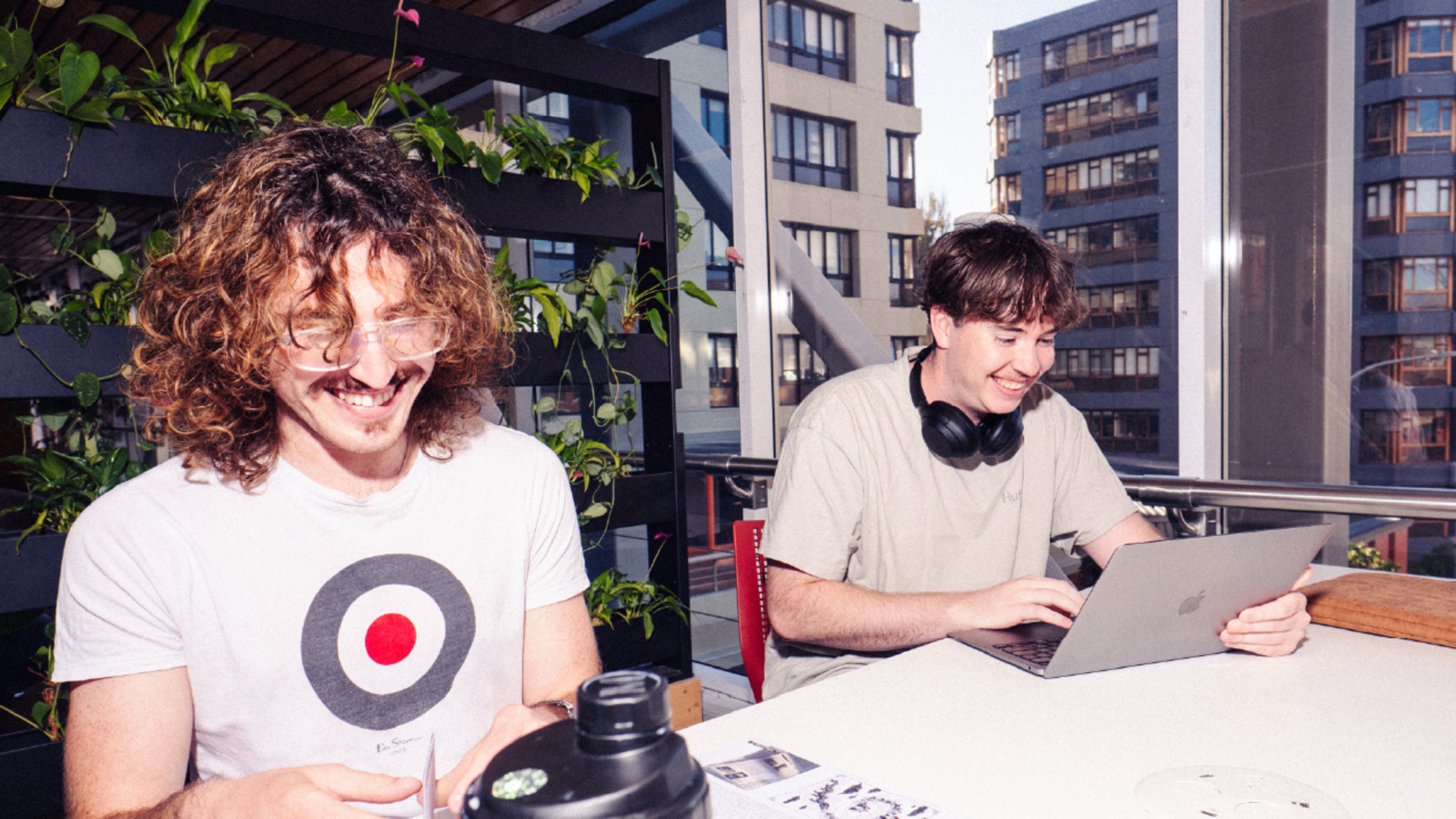 Two people sit at a table indoors with a laptop, papers, and a water bottle; buildings are visible through large windows behind them.