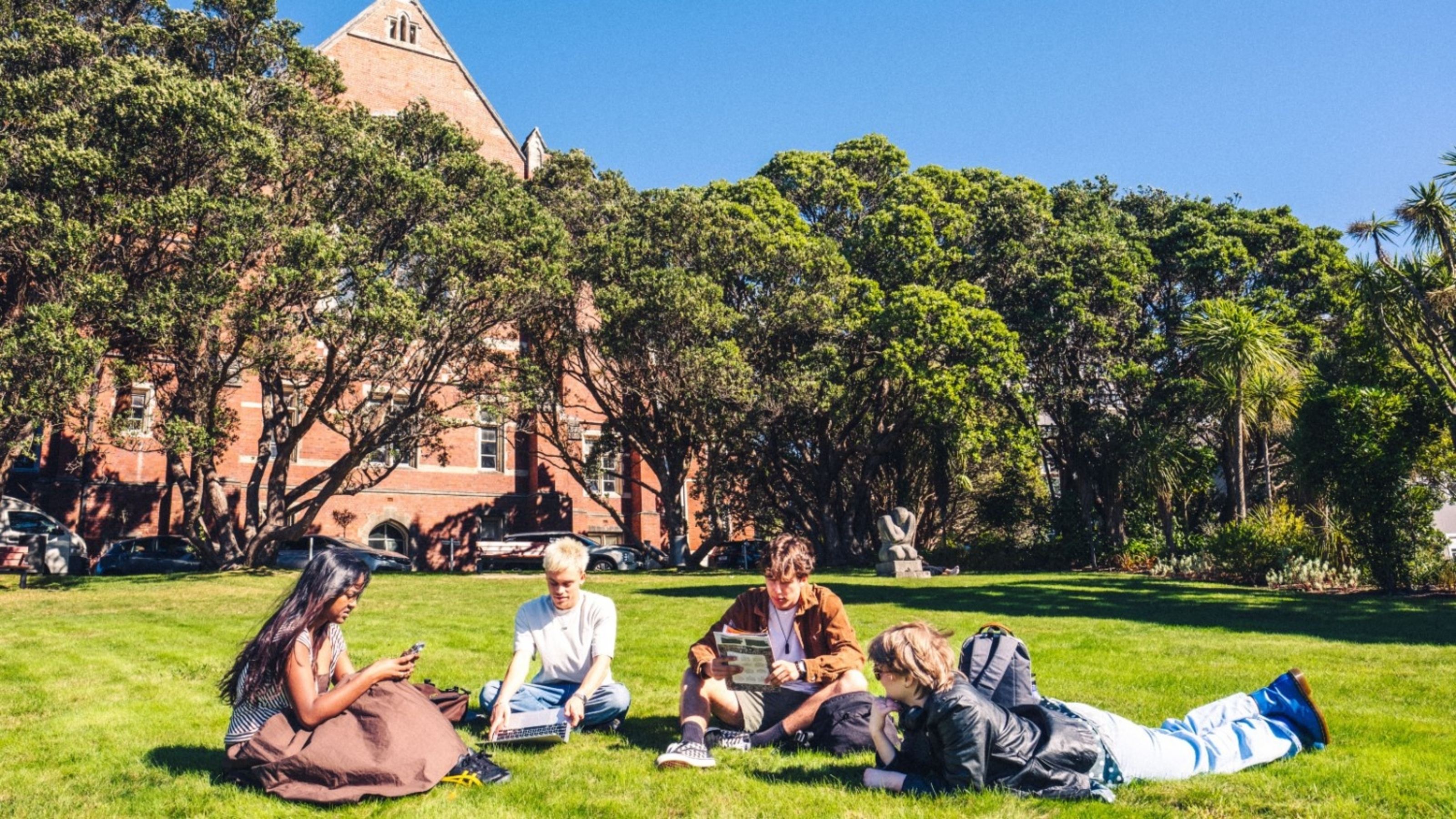 Two female and two male students are sitting or lying on the Hunter lawn at Kelburn campus on a sunny day. One student is on their laptop, another student is on their phone, and another student is reading a magazine.