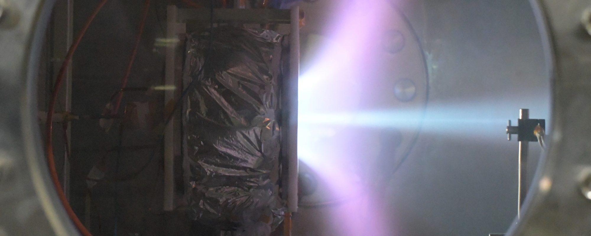 Looking through a vacuum chamber window, a plasma thruster is being tested