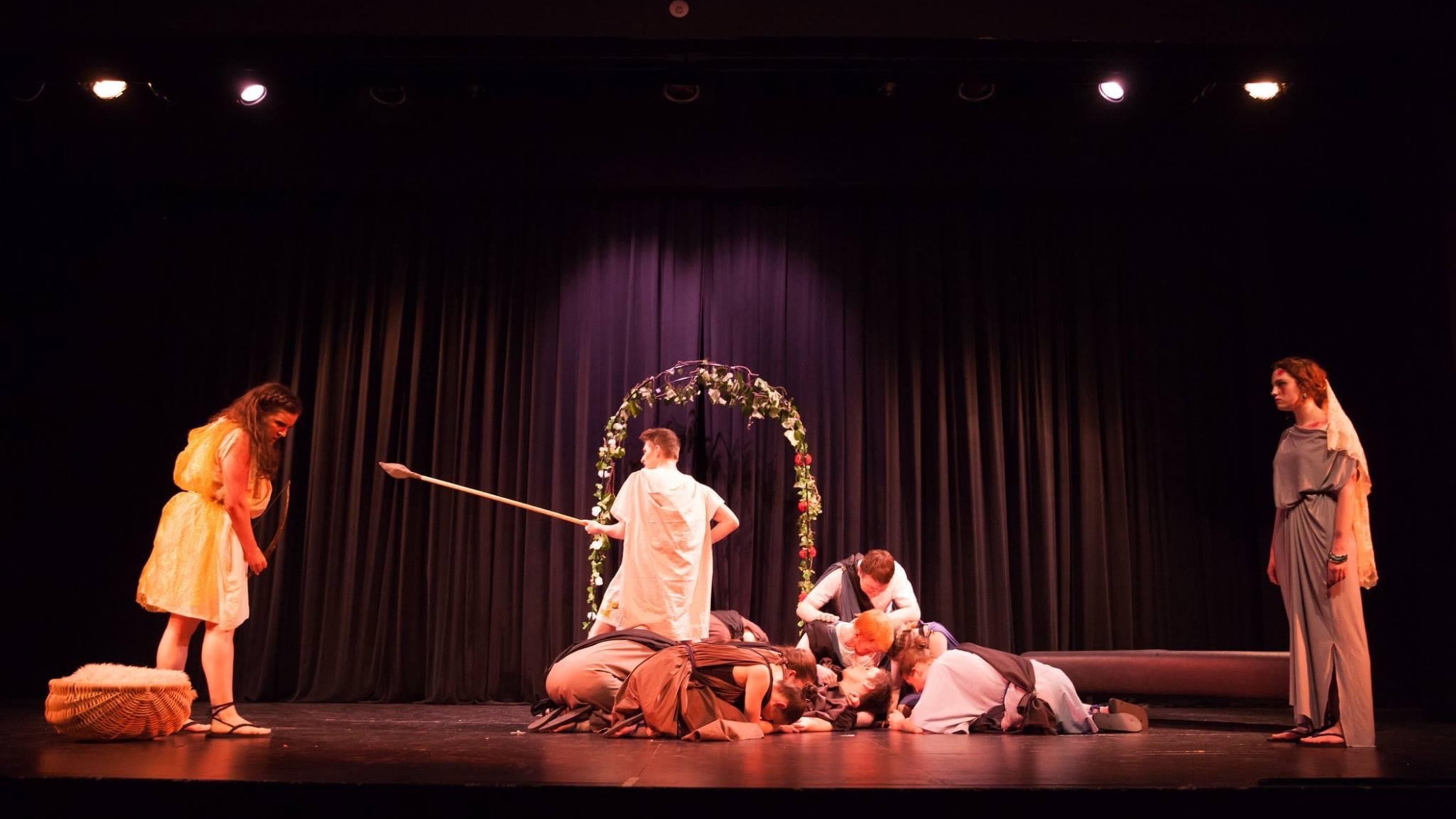 Final scene of performance, with actor amassed in the middle, man in white toga pointing spear against woman in toga.