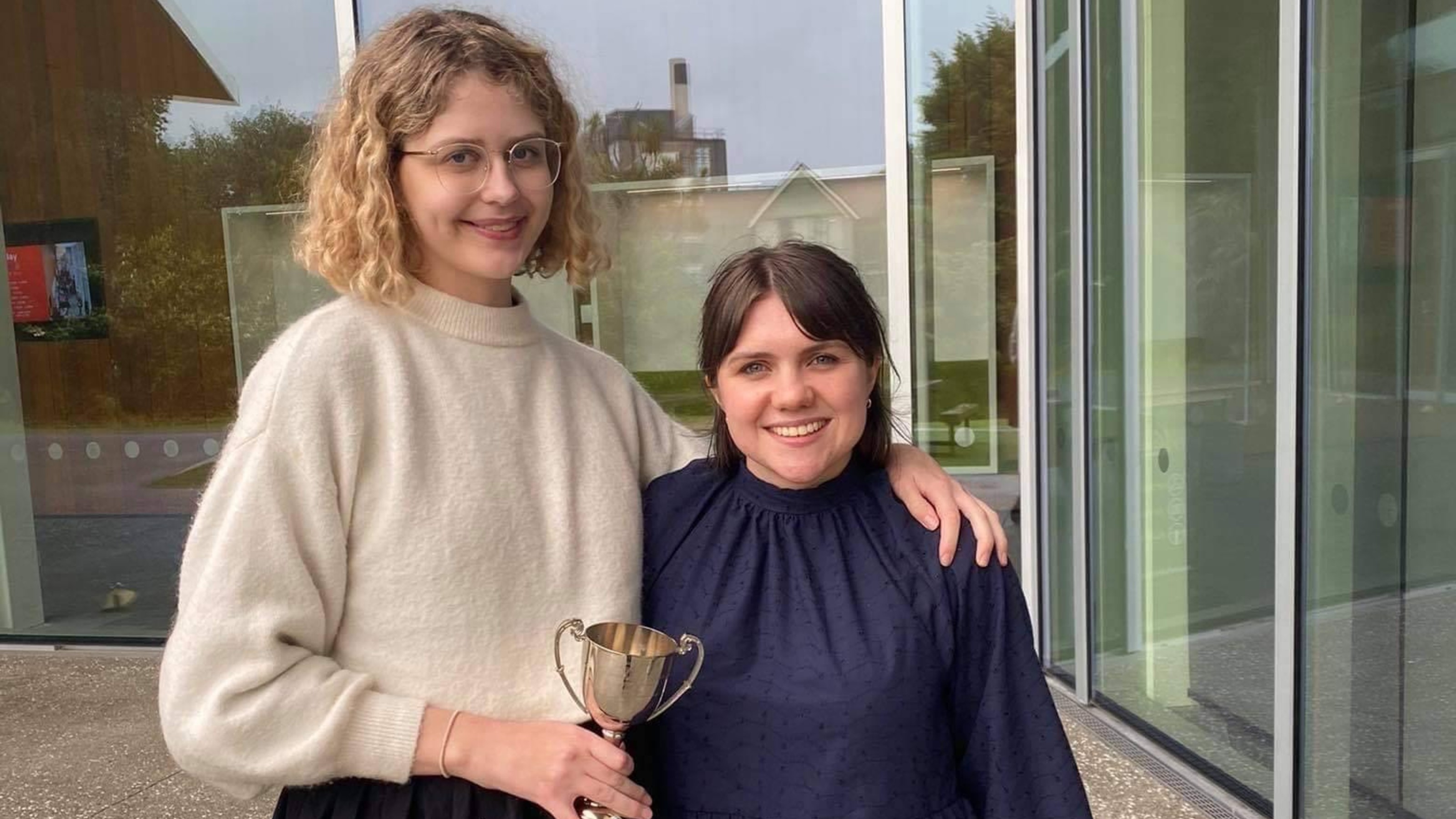 Two winners of girls debating competition, standing before glass building, holding cup.