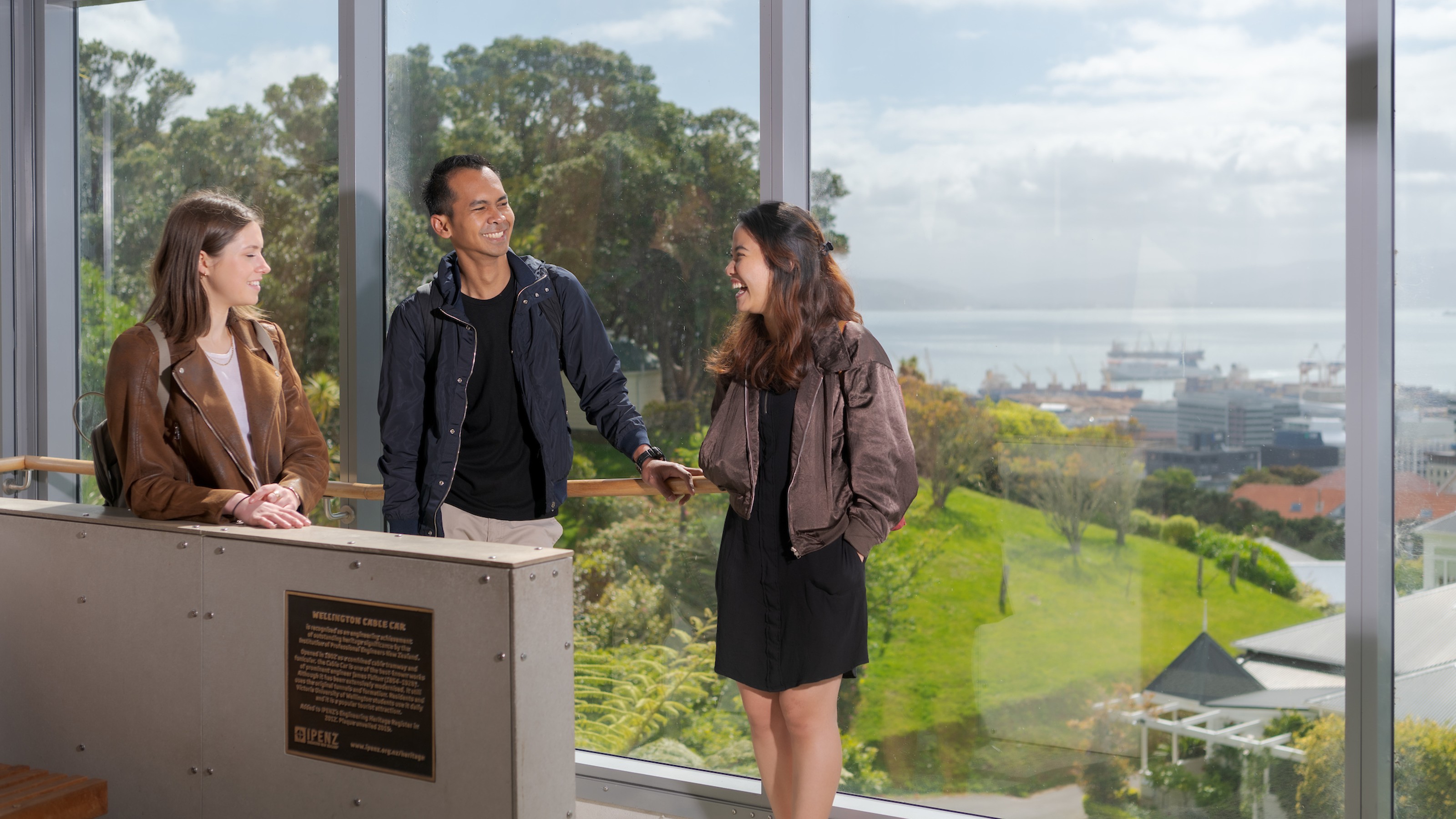 Three people chatting at the top of the Wellington Cable Car, with a panoramic view of the city in the background.