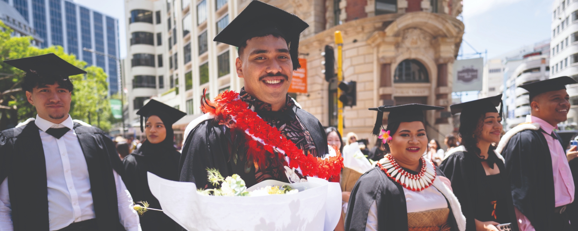 Graduate in cap and gown holding flowers leads a group of graduates walking through a city street during a procession.