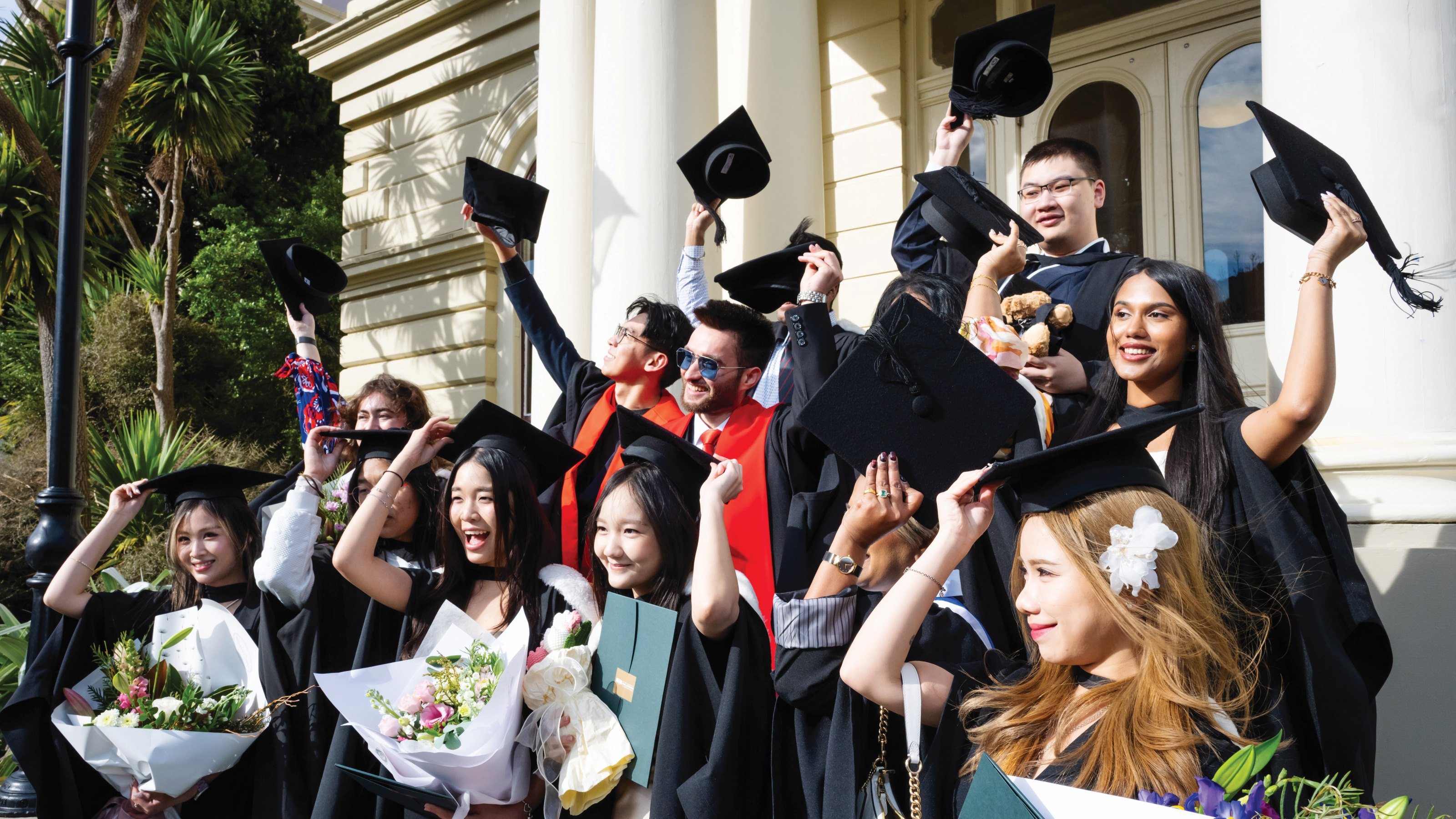 Graduates in academic gowns celebrate outside a building, raising caps and holding bouquets.