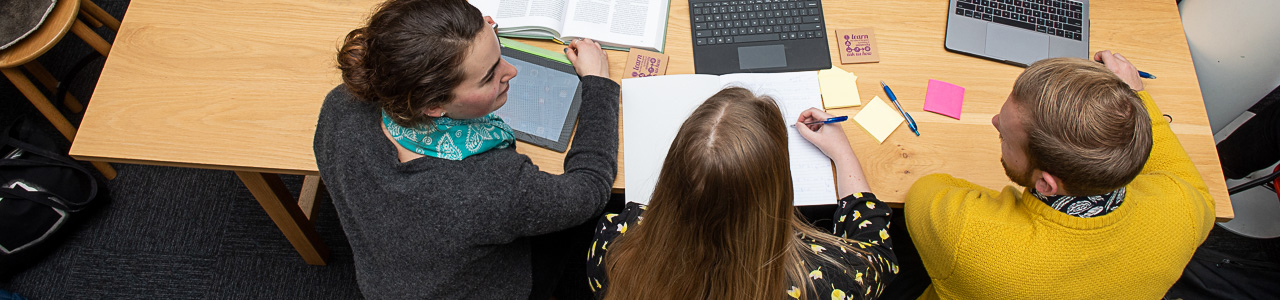 A top-down view of three students working collaboratively at a table, using laptops, a tablet and a notebook.