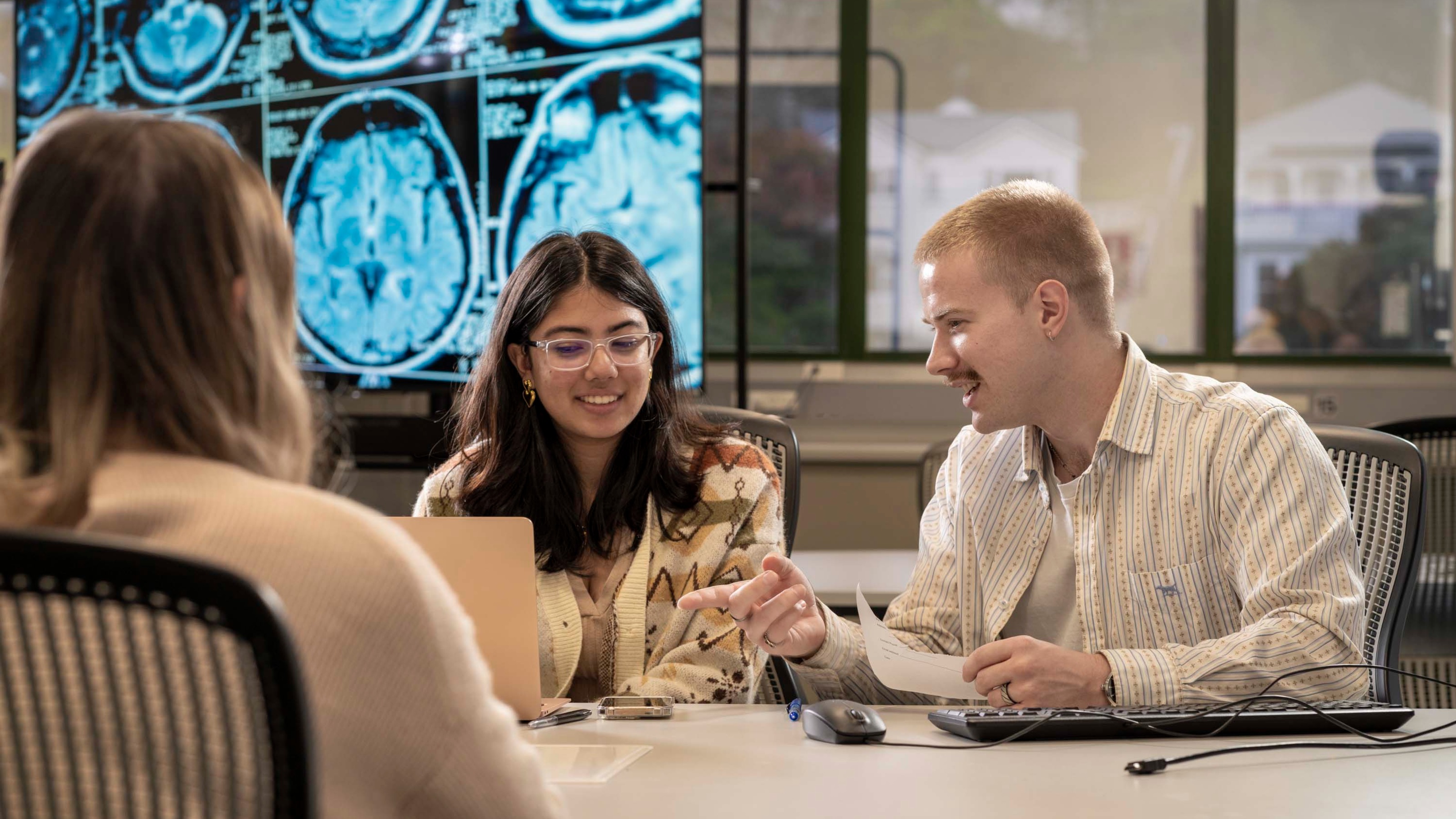 Three students talking with computer equipment in front of a lightbox displaying scans of brains.