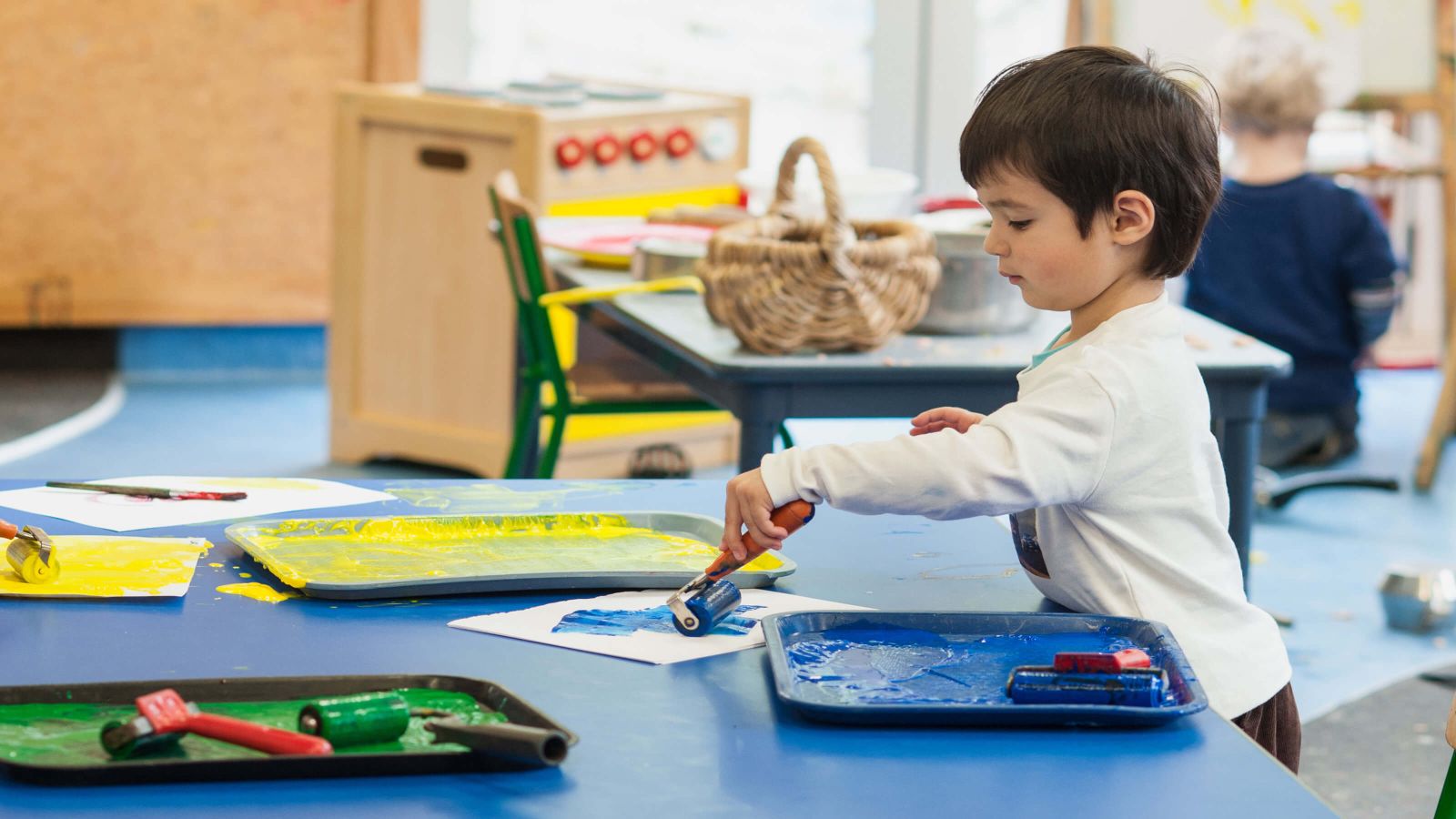 Paint Time – A young boy in a children’s activity room, stands at a short desk with trays of yellow, green and blue paint. He holds a paint roller of blue paint and creates a masterpiece on white paper, all the while, looking quite pleased with his work.