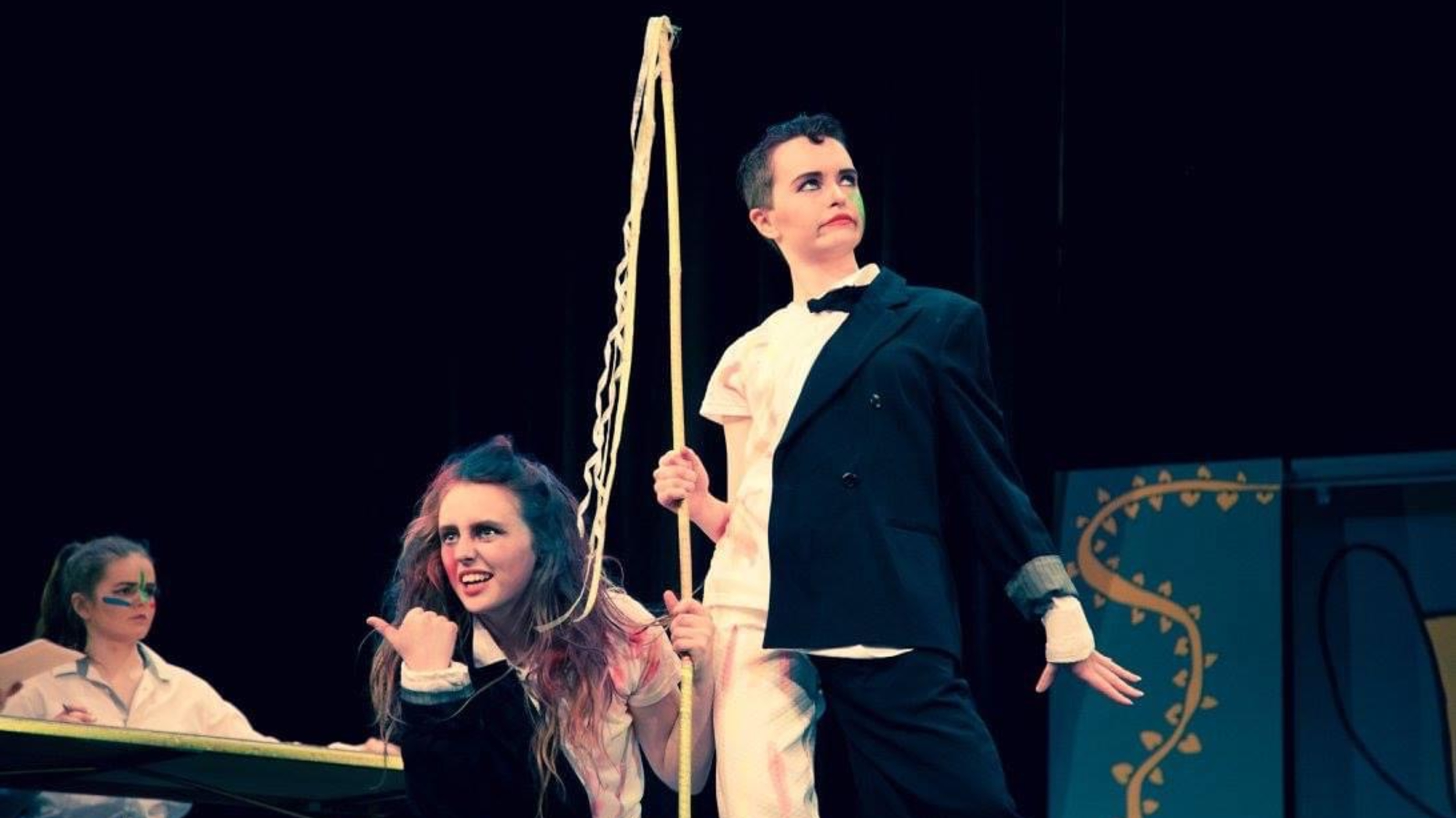 Still from play: three actors with face paint, one sitting at a desk, one facing the public, one in half costume half t-shirt looking in the distance.