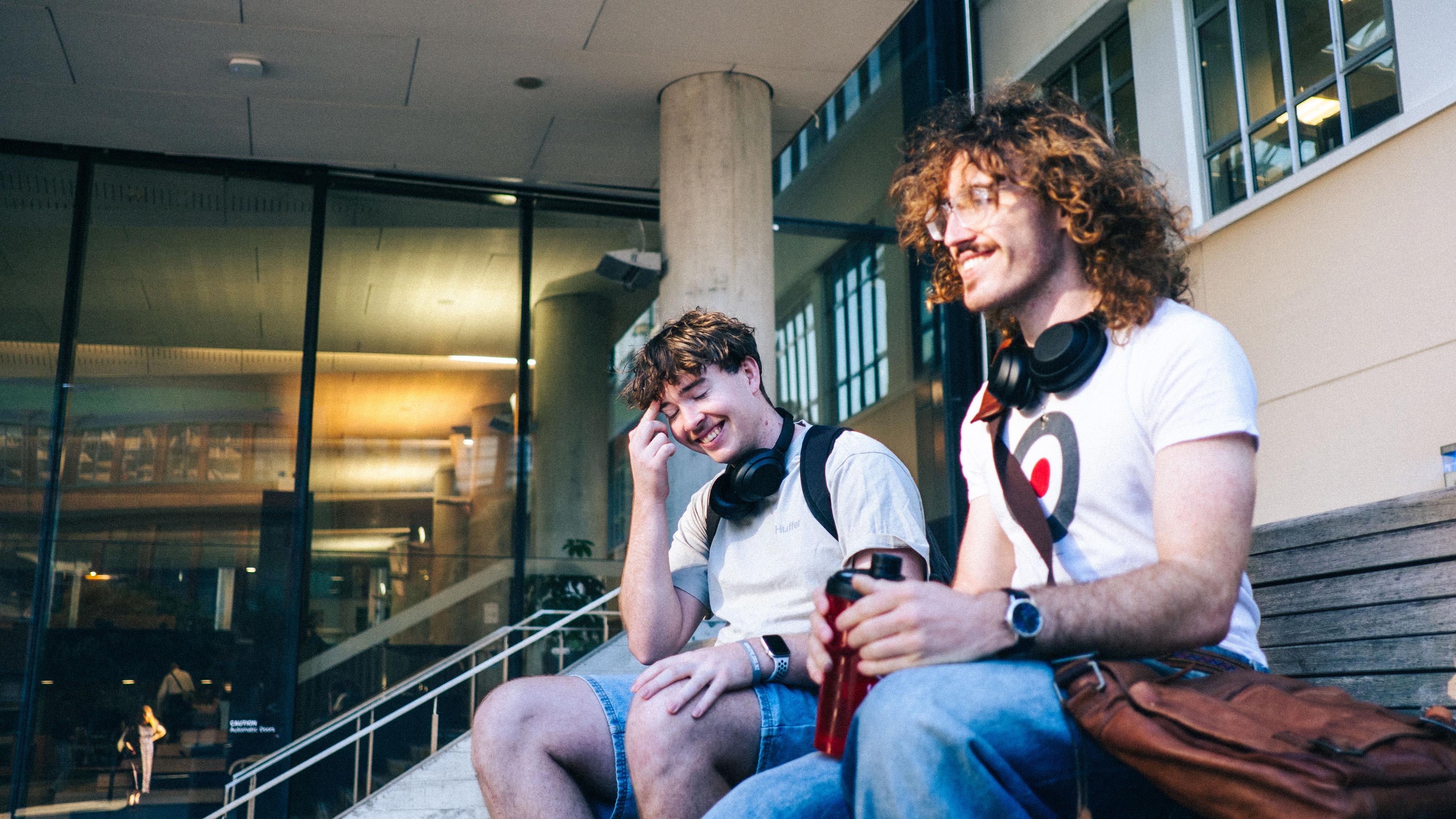 Two students sitting on stairs on campus, with headphones around their necks