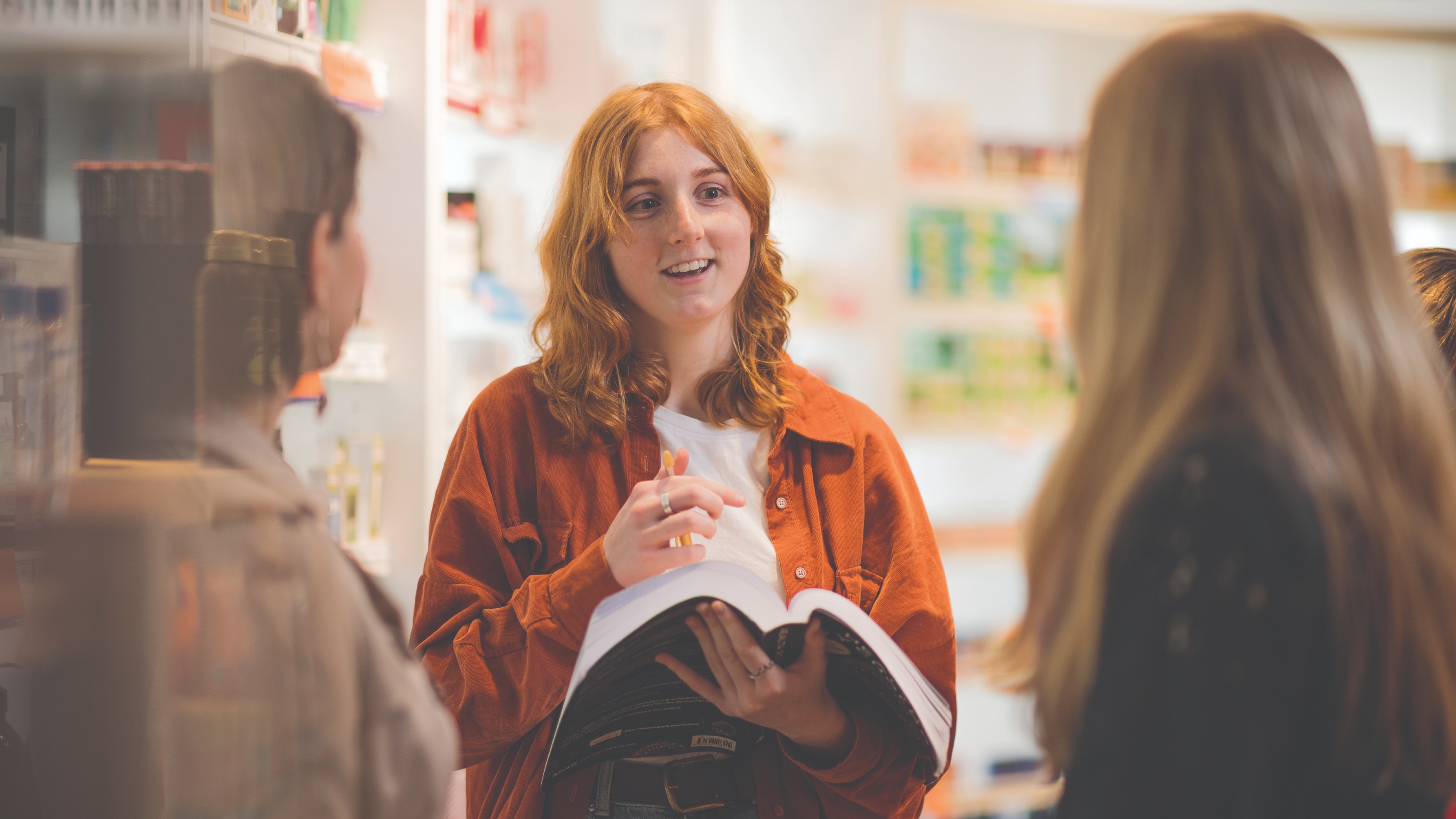 A redhaired woman holding a book, flanked by two other students in a library setting.