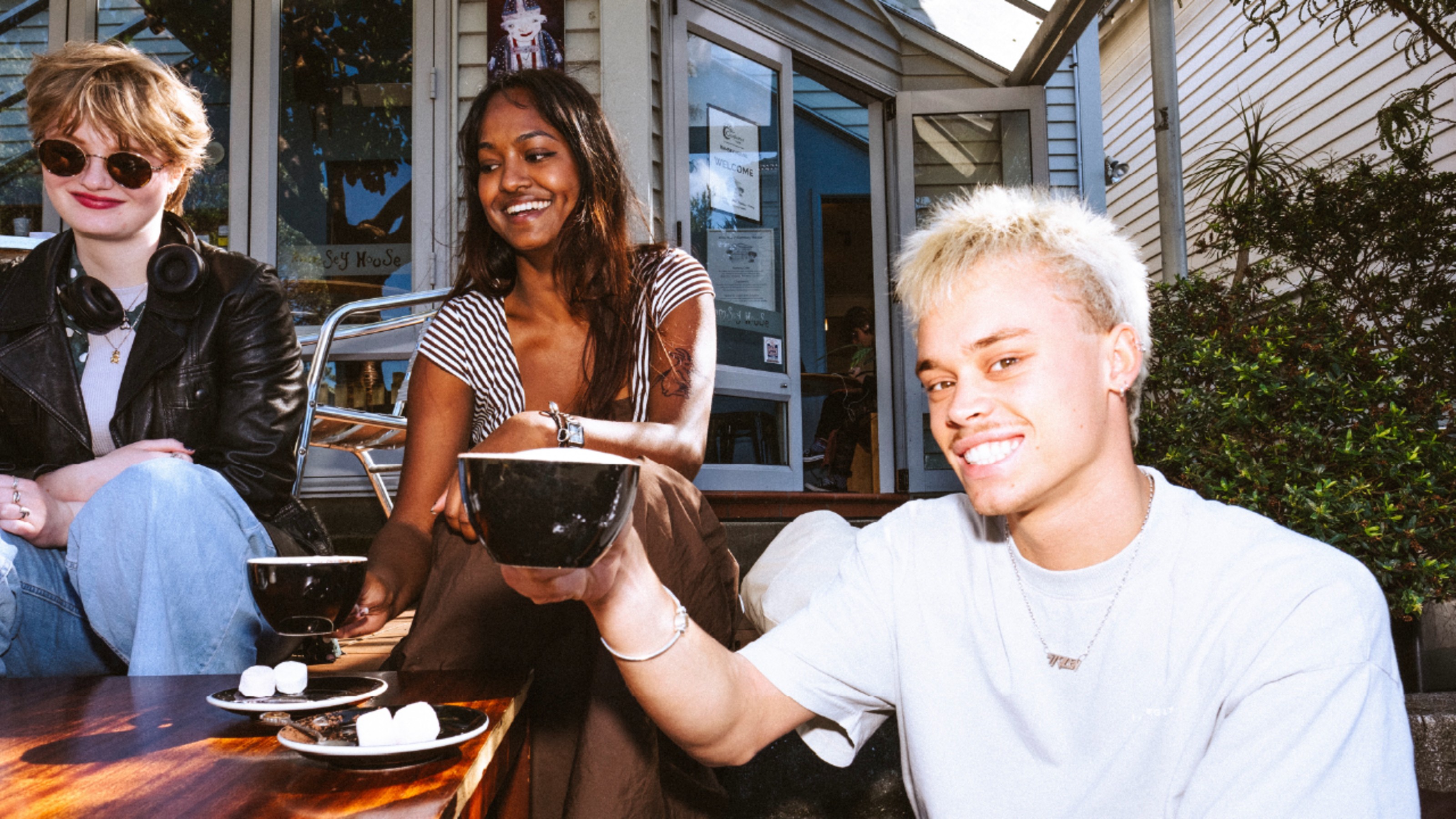 Three people sit outside a café holding black cups, with plates of marshmallows on the table in front of them.