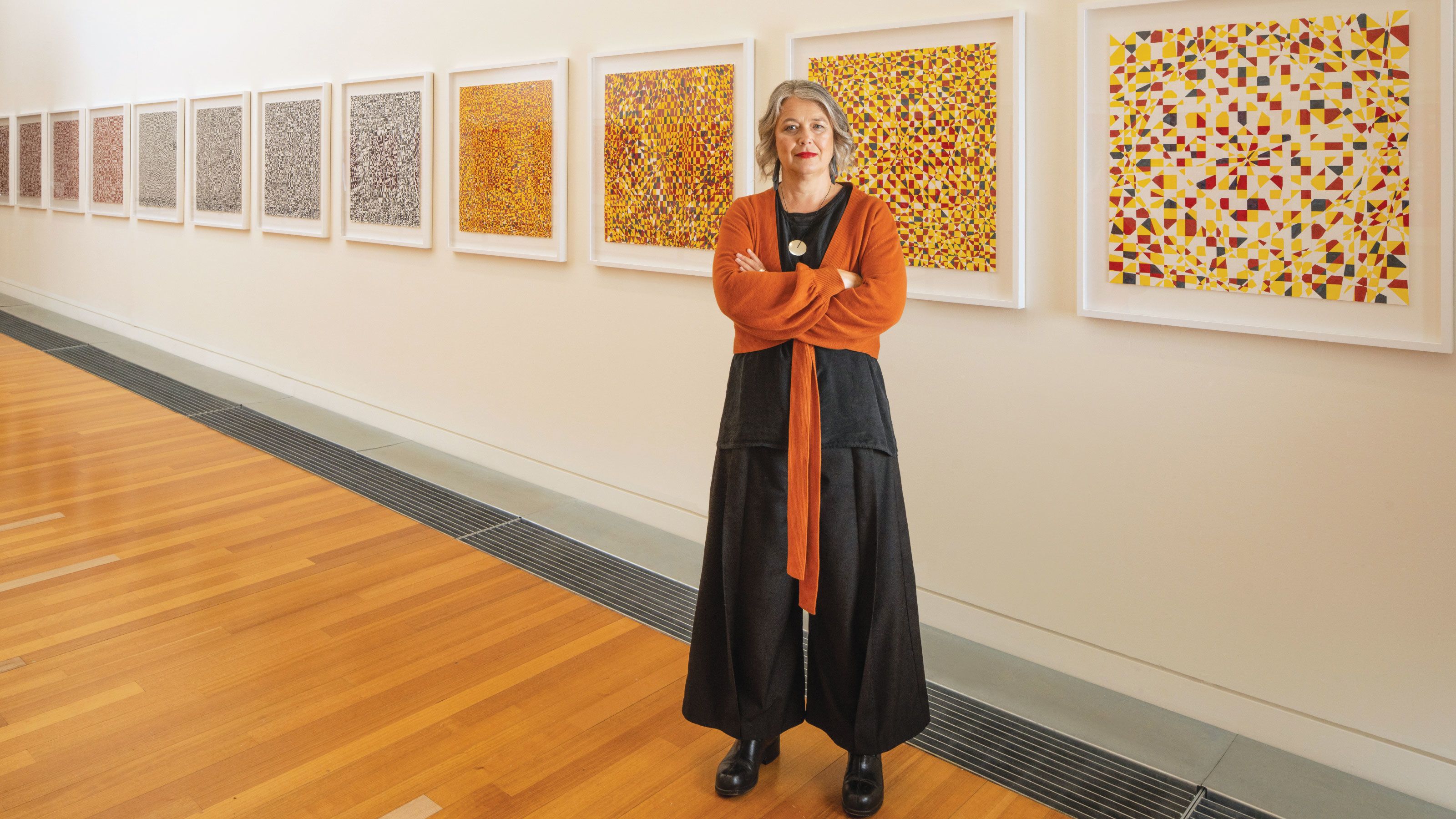 Associate Professor Su Ballard stands in front of a wall hung with art works in the Adam Art Gallery.