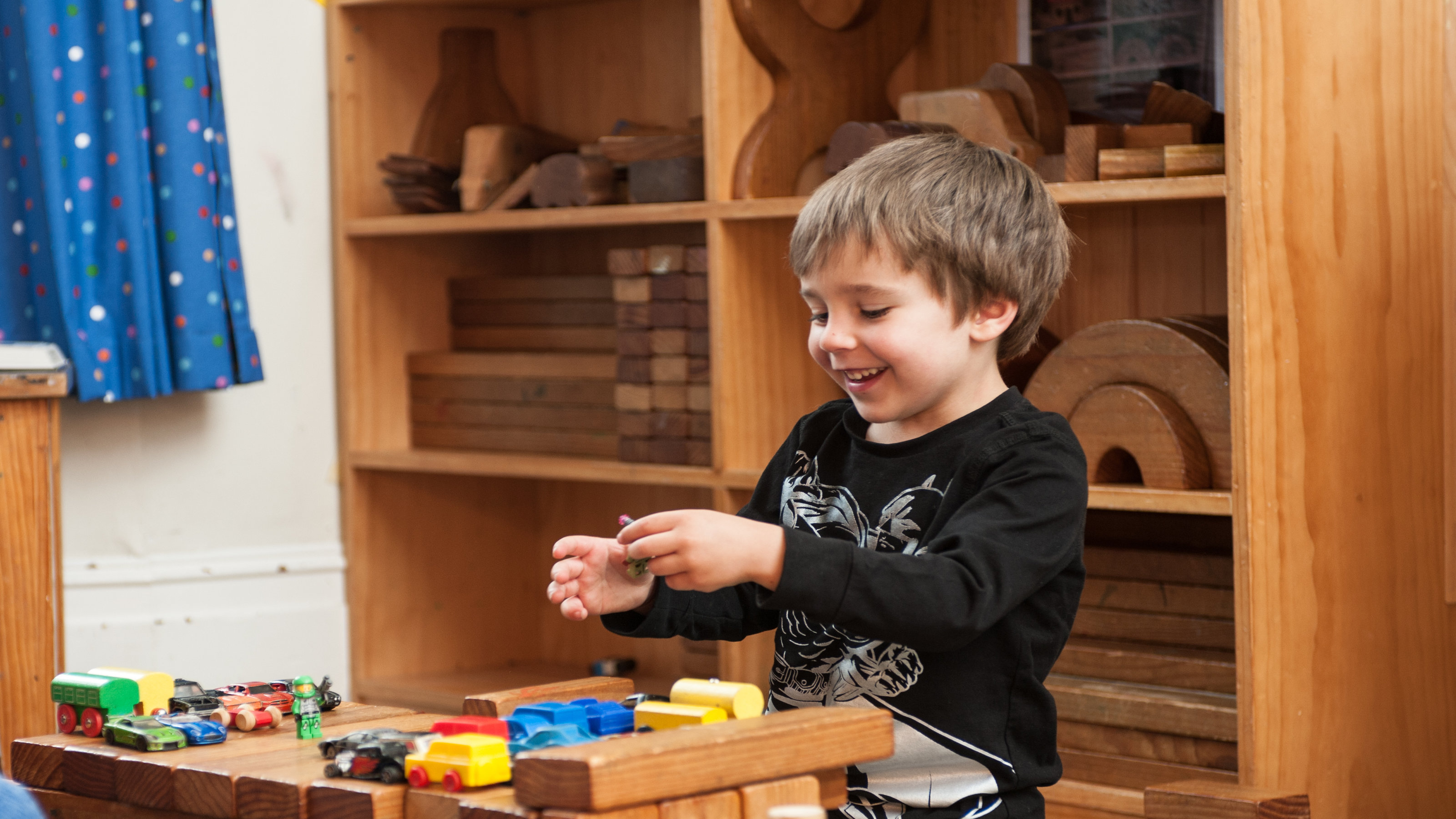 Block Time – A young boy in a children’s activity room, smiles while playing with a lego character at a group of blocks with toy cards and another lego character are set up, for an imaginary game.