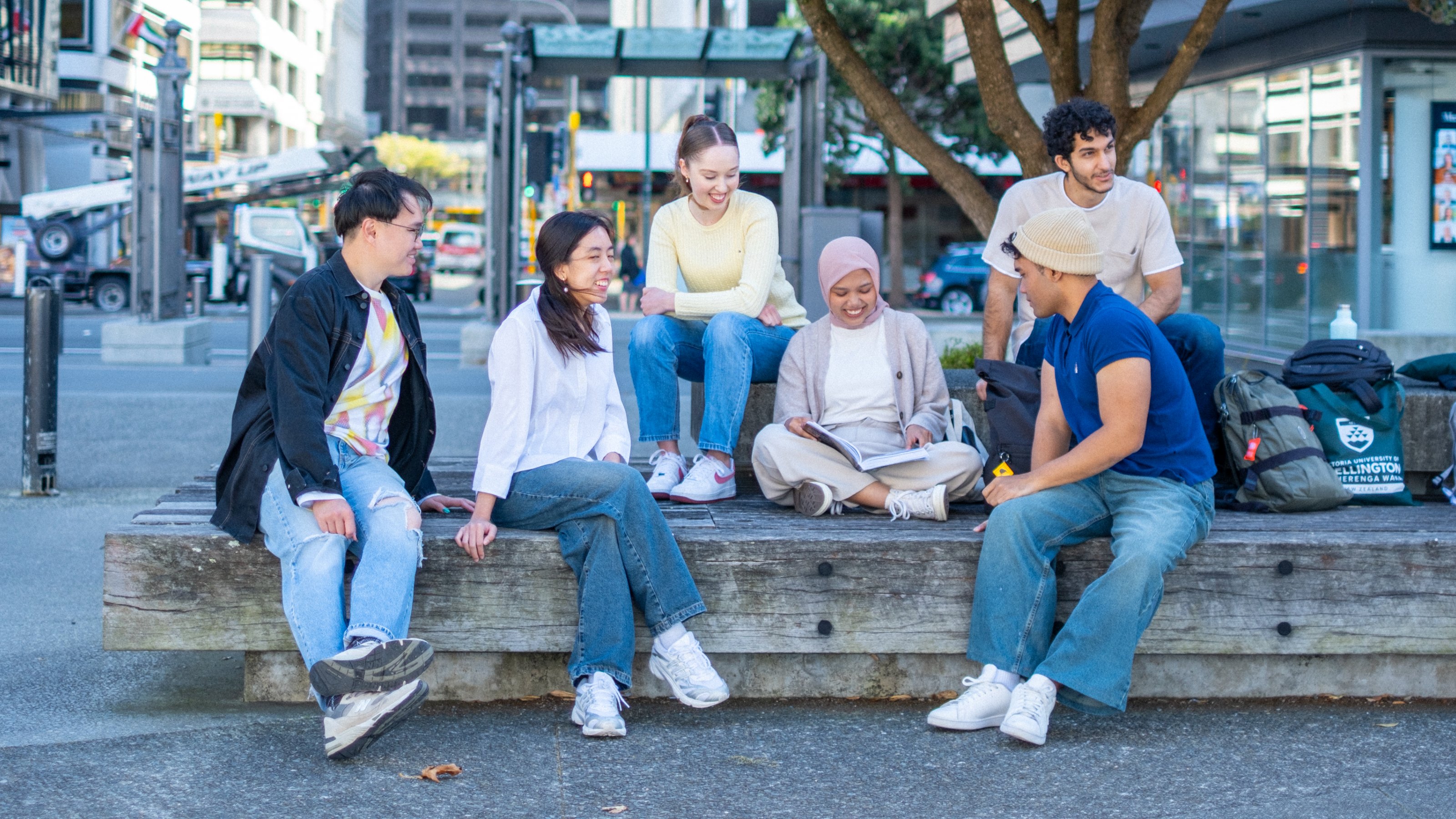 Six students of apparently different nationalities sit on outdoor bench in downtown location