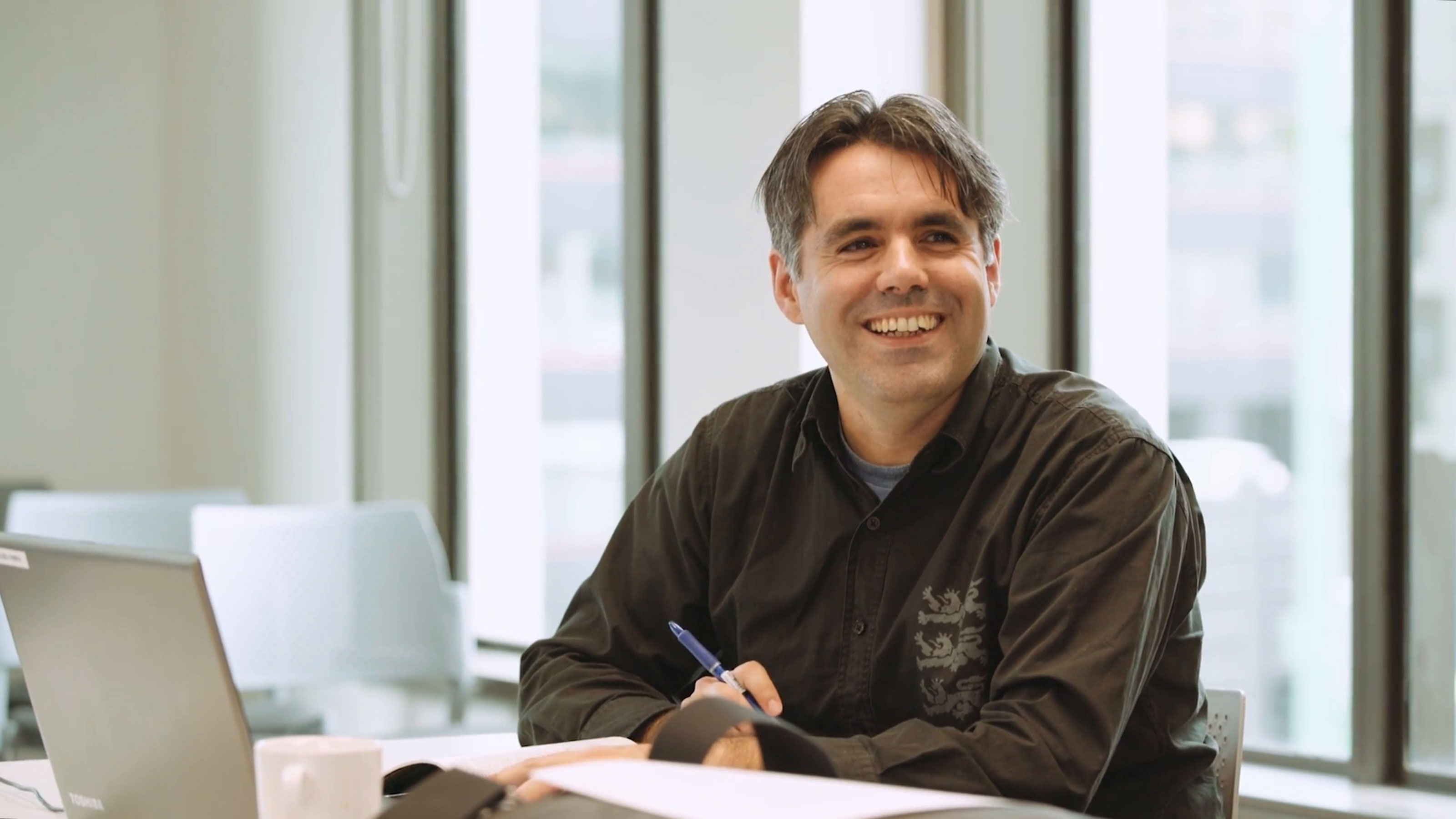 A man sits smiling at a desk with a laptop and paperwork in an office.