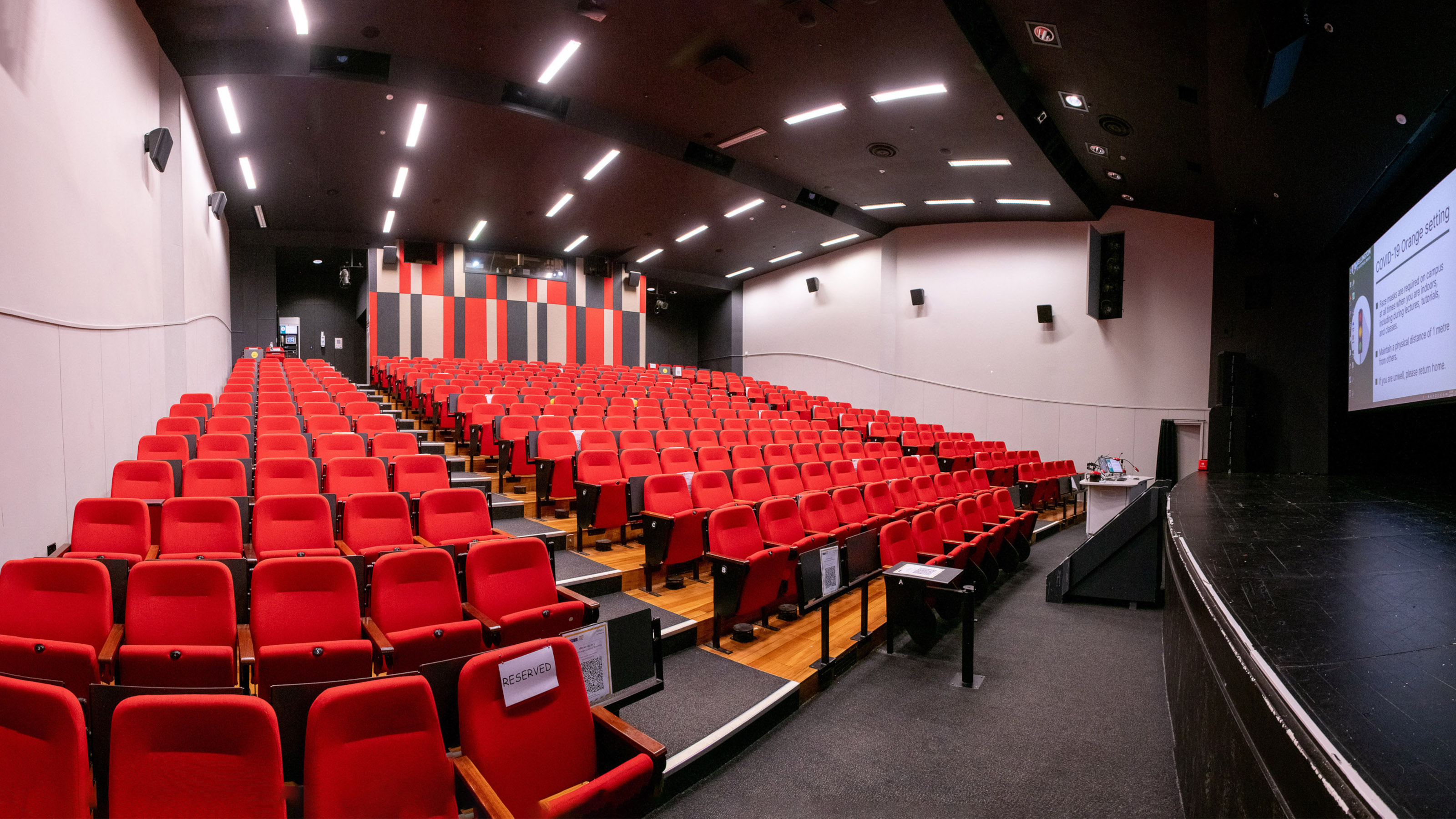 View of Memorial Theatre from bottom left of auditorium. Shows bank of tiered red coloured seating, computer screens and microphones on rostrum at the front of theatre, and large screens at front of theatre.
