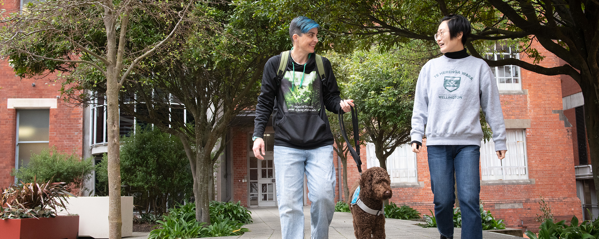 Two people walking out of the Hunter building lecture theatres into the courtyard. One has a support dog, the other is wearing a Victoria University of Wellington sweatshirt.