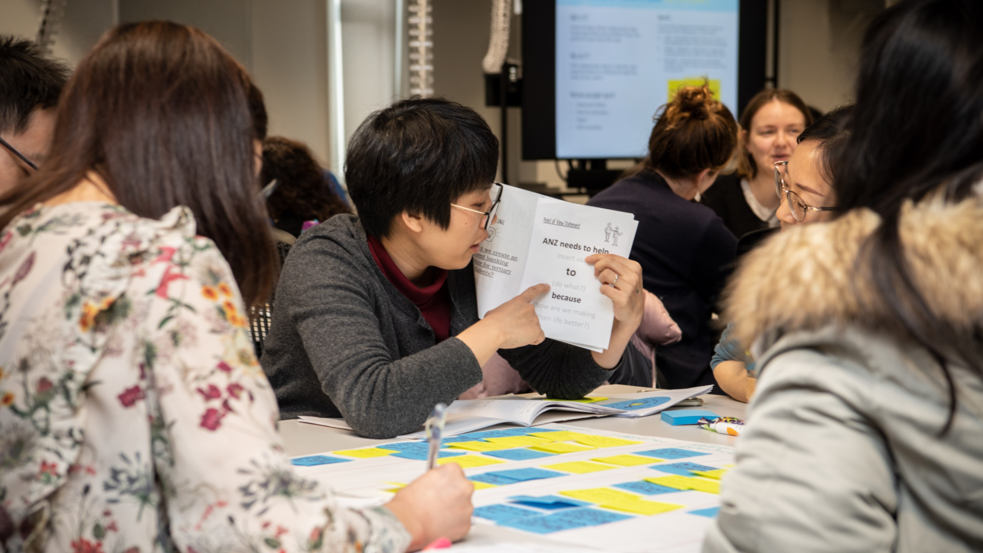 workshop 5 Students sitting around a table with yellow and blue post-it notes laid out in rows on the table. One student is holding up an open book and pointing to the page.