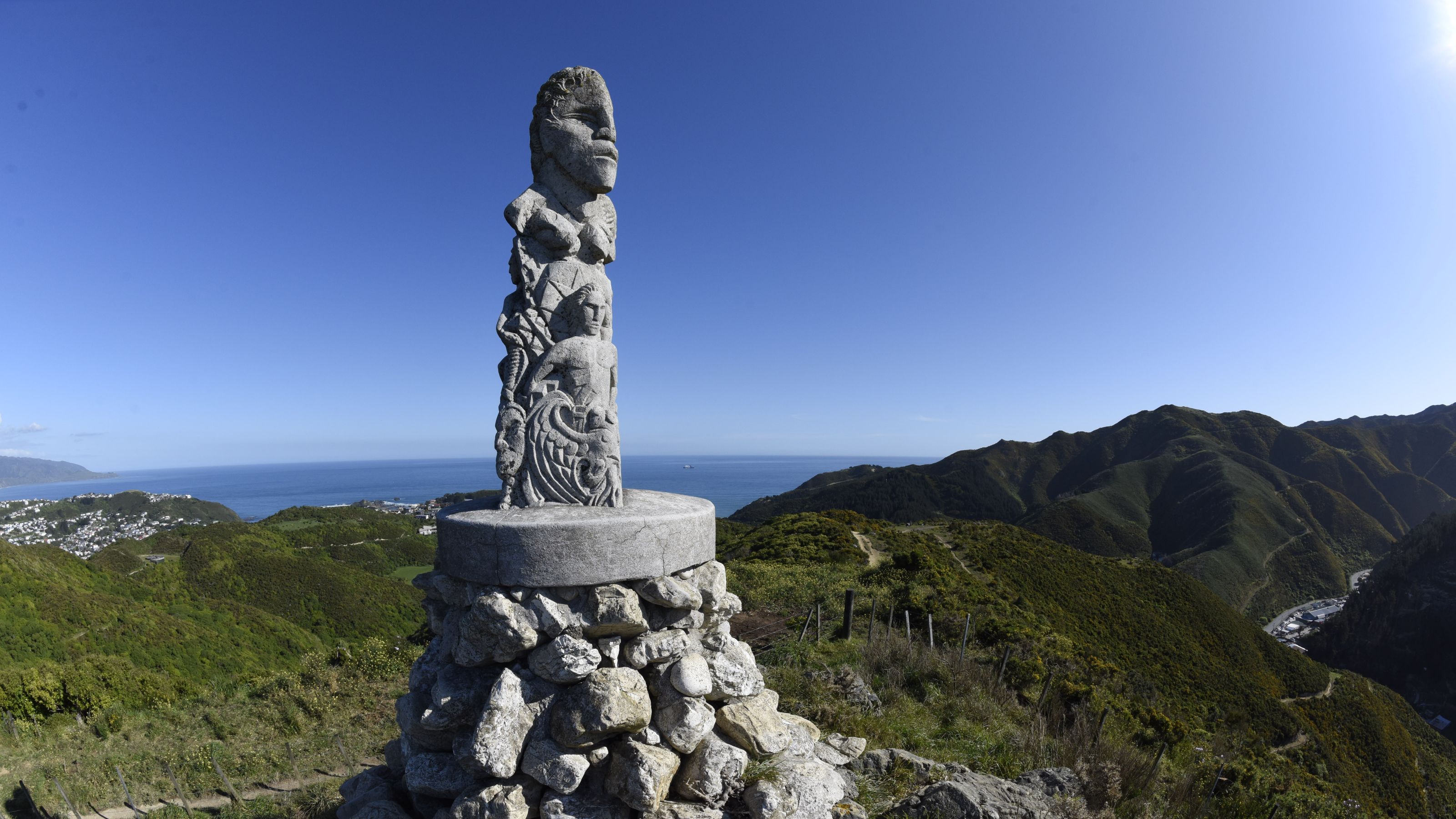 Stone sculpture of Māori design at the top of a ridge of land, overlooking the horizon with blue sky.
