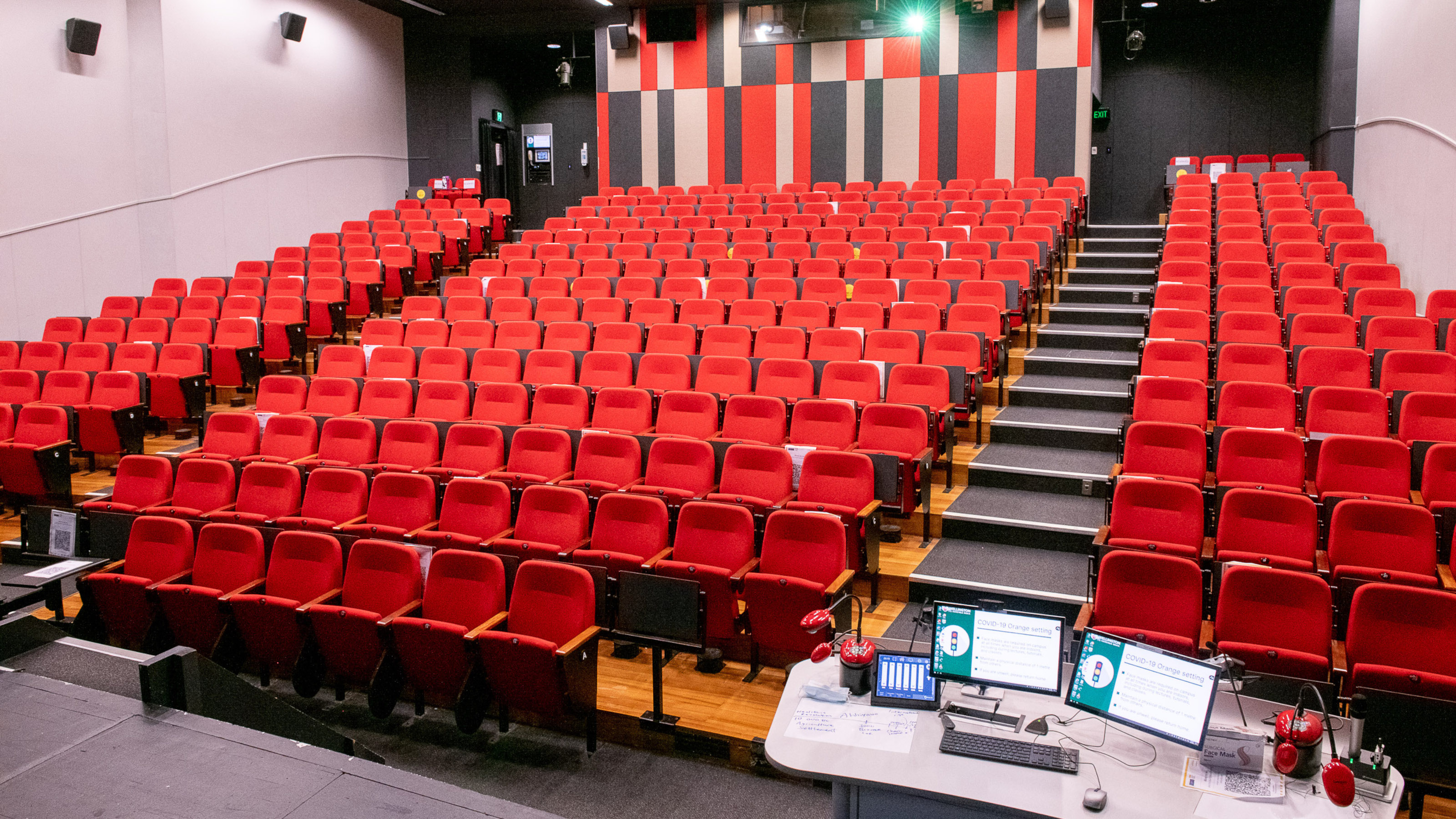 View of Memorial Theatre from bottom right of auditorium. Shows bank of tiered red coloured seating and computer screens and microphones on rostrum at the front of theatre.