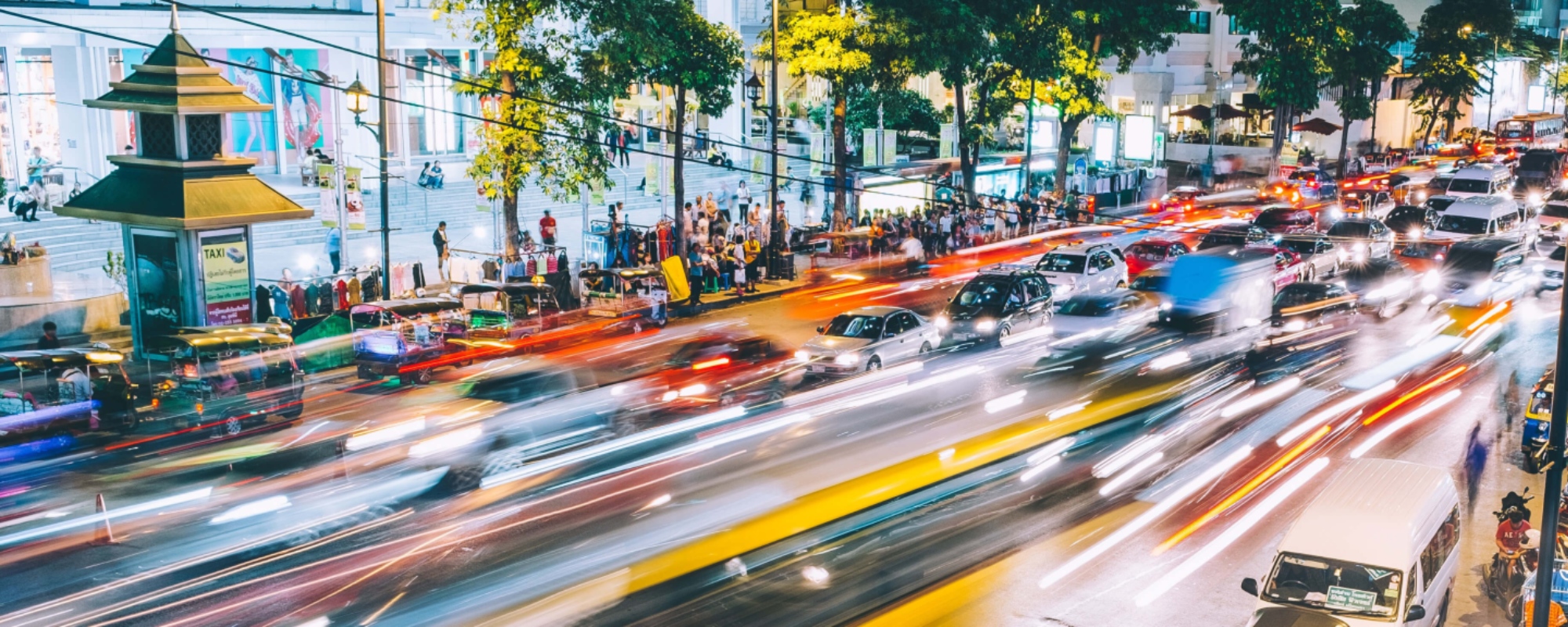 Busy urban street at night with vibrant light trails from moving vehicles, parked cars, and motorbikes.