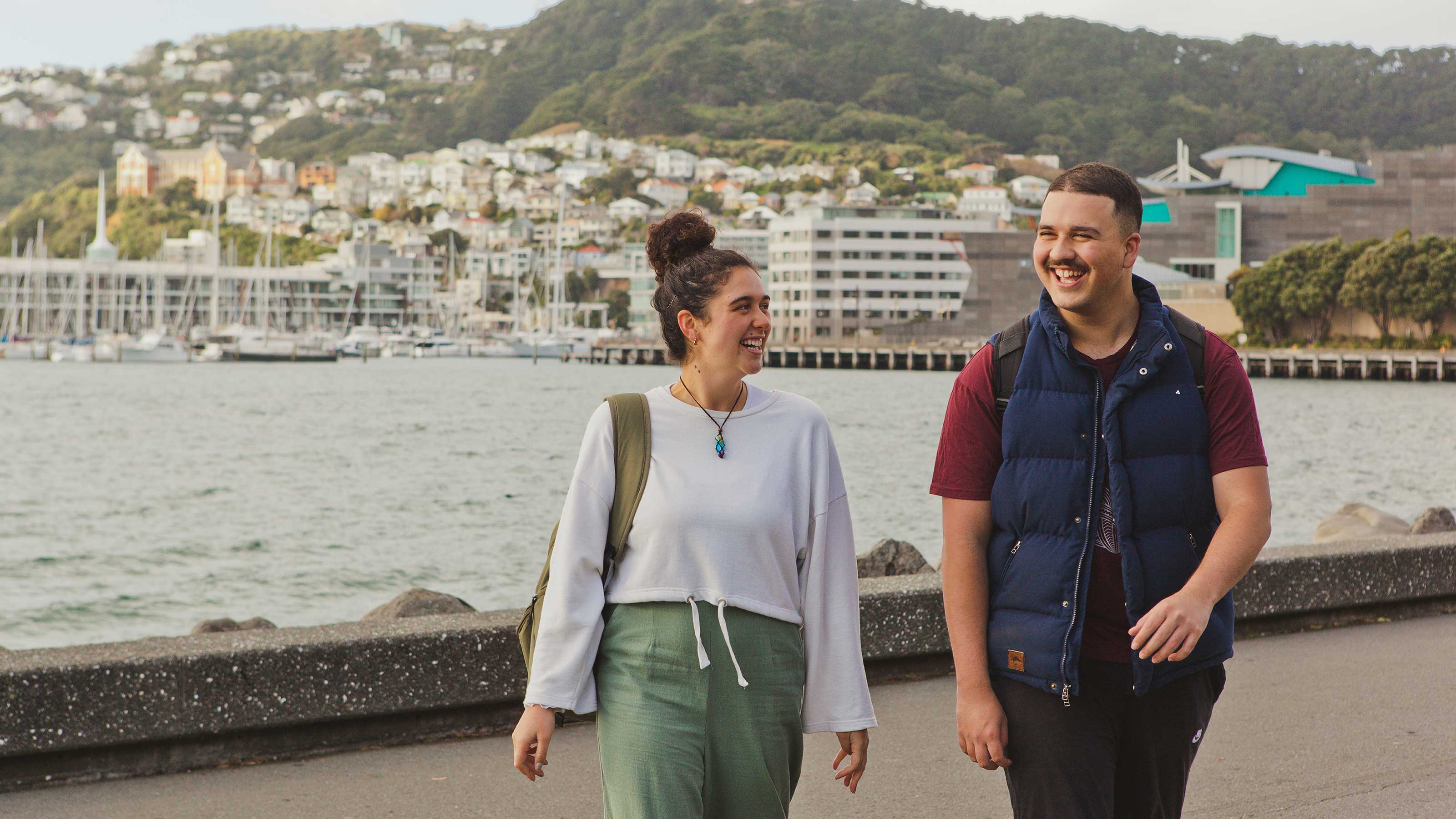 Two students walking along the waterfront with a picturesque harbor in the background, featuring a backdrop of buildings and boats docked along the shoreline.