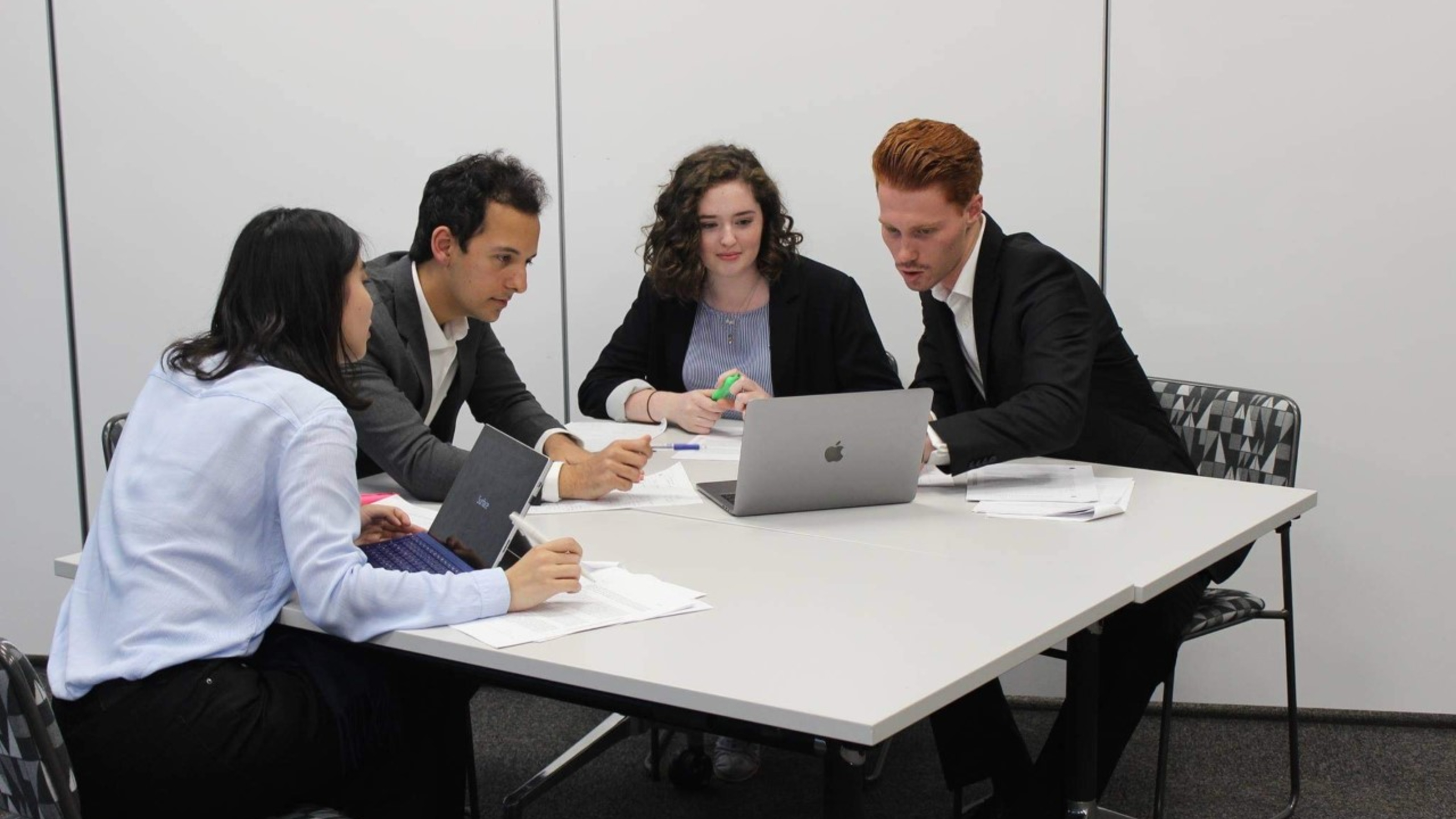 Four students working together at desk, with laptops and papers.