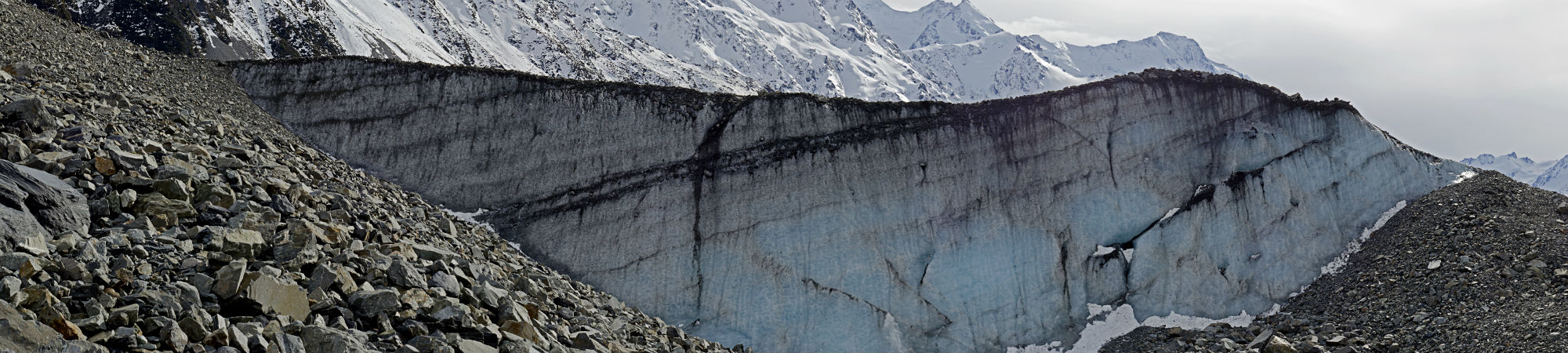 Steep snow-capped mountains with a retreating glacier showing ice strata and a scree slope with loose rocks in the foreground.