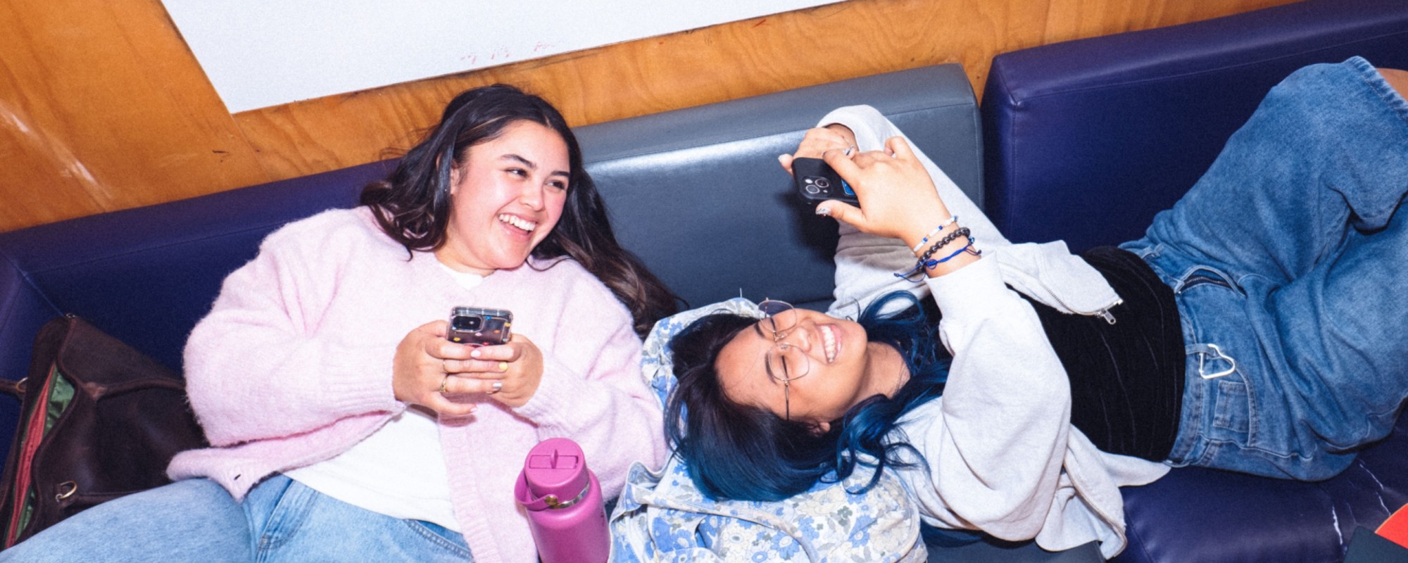 Two students relaxing on a couch sharing something on their phones.