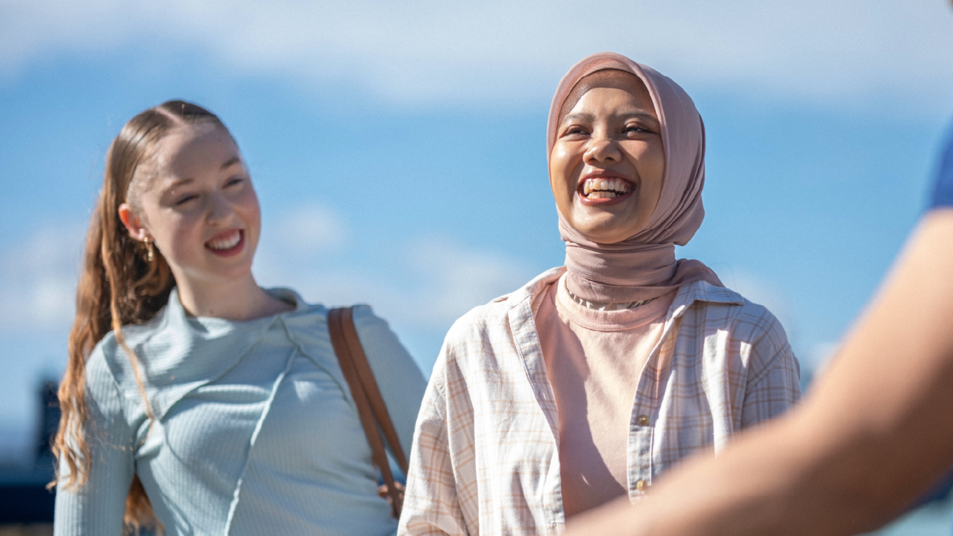 Two students stand outdoors under a clear blue sky; one wears a light blue top, the other a pink hijab and an open overshirt.