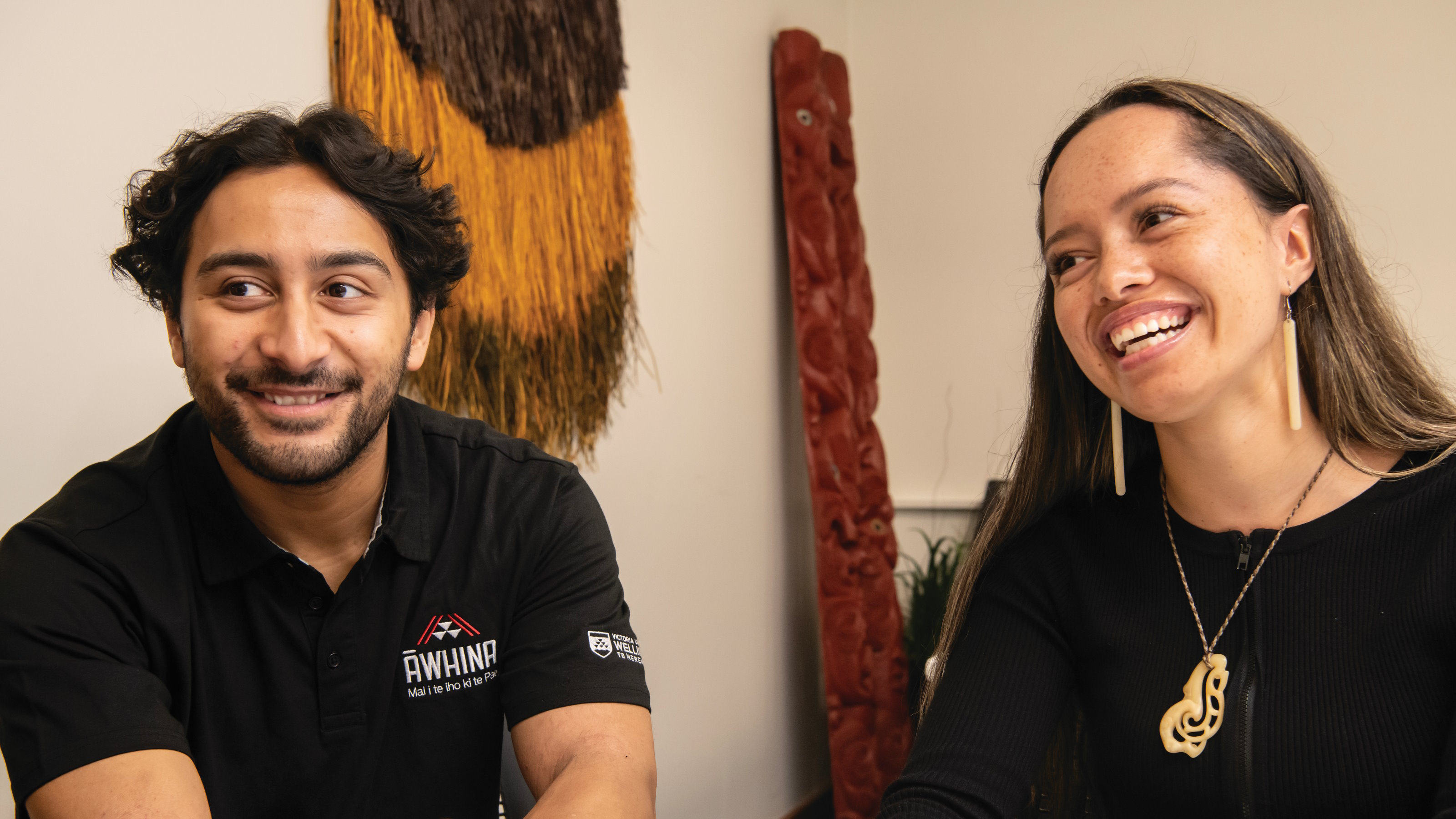 A young Māori man wearing a black shirt with an Awhina logo and a young Māori woman, both laughing.