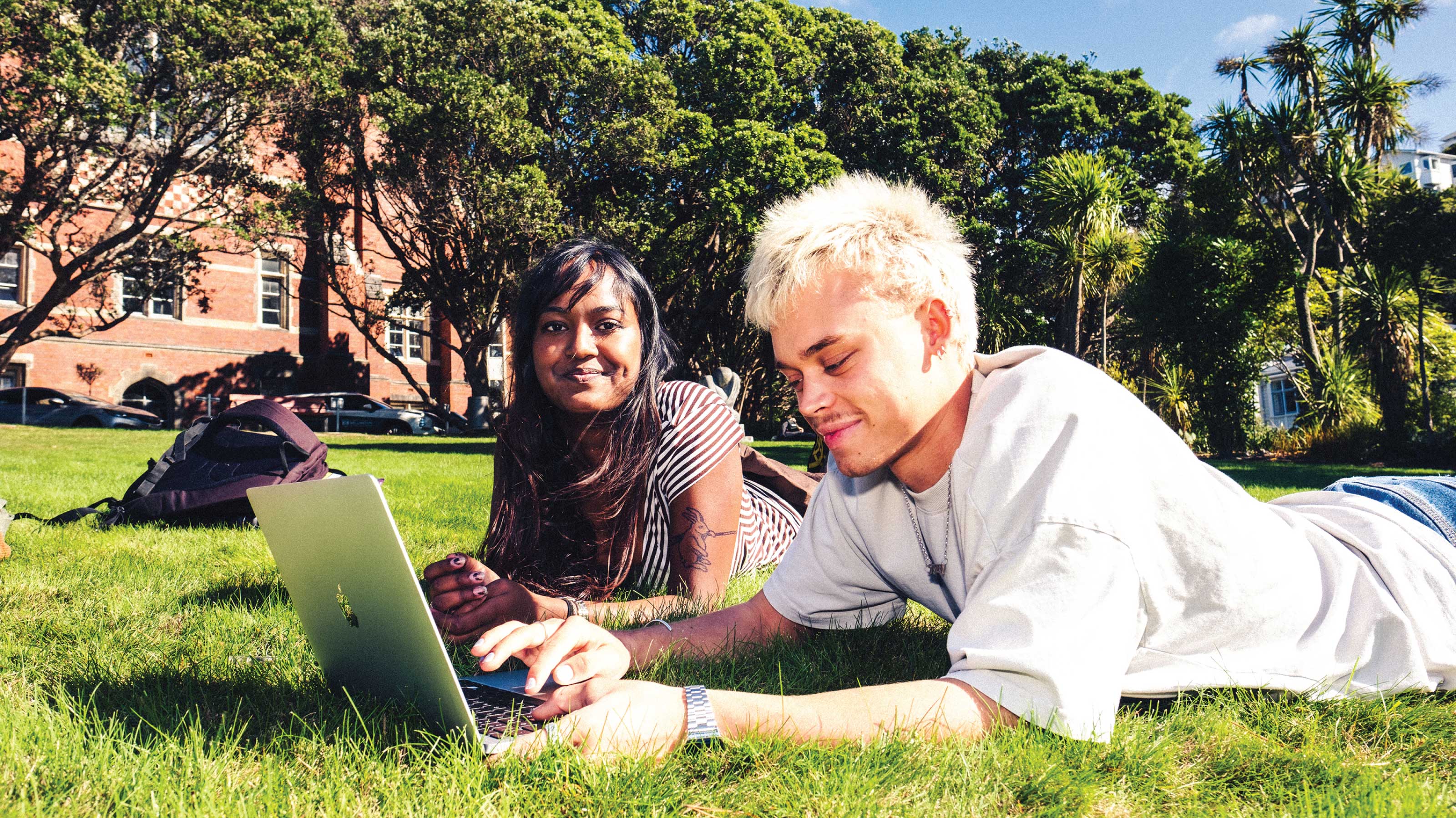 2 students lying on their front on grass. One is looking at the camera, the other is looking at an open laptop screen