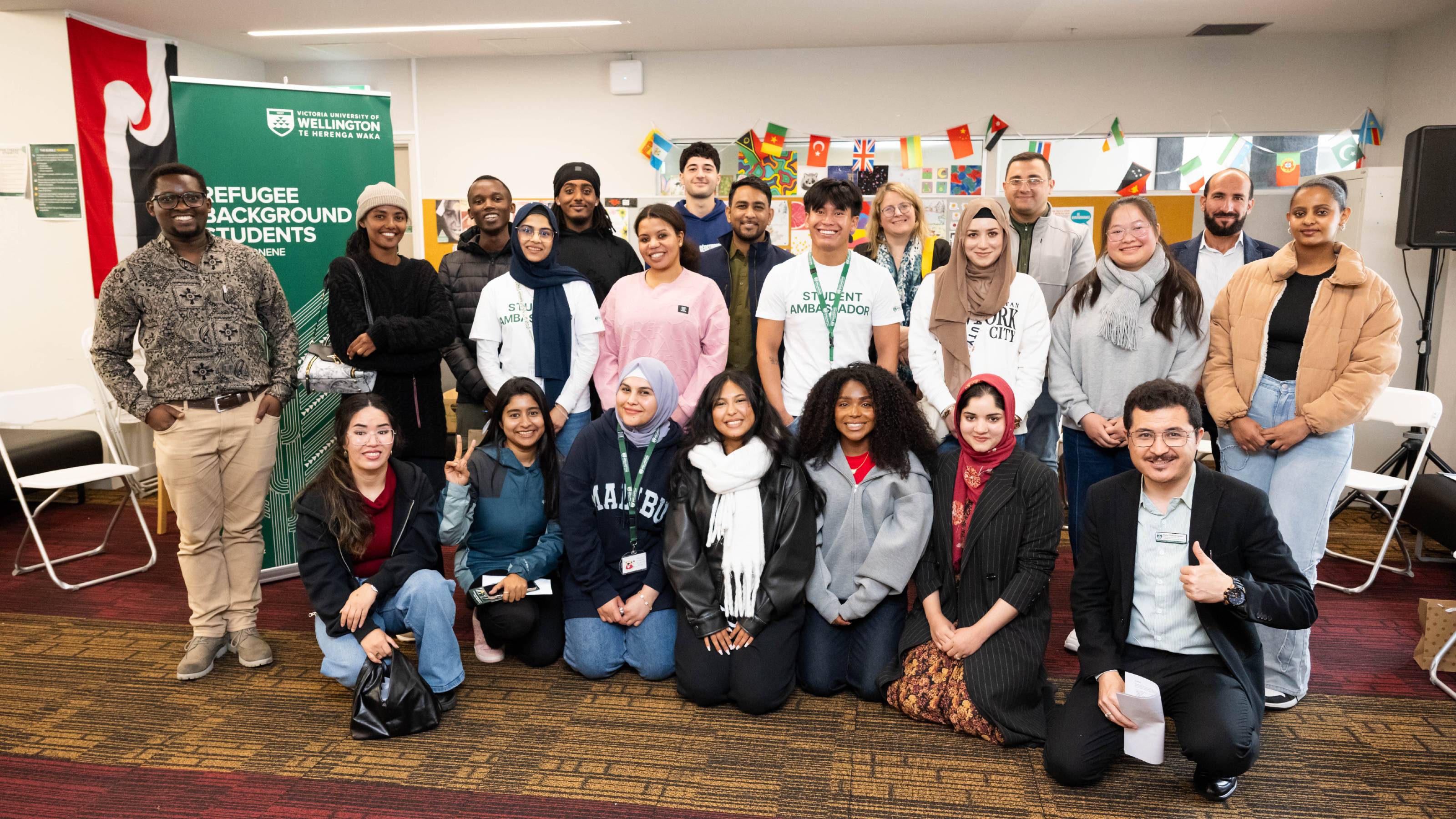 A group of 20 people, students and staff smiling at an event in The Bubble—Kelburn Edition, with a banner that reads Refugee-background students.