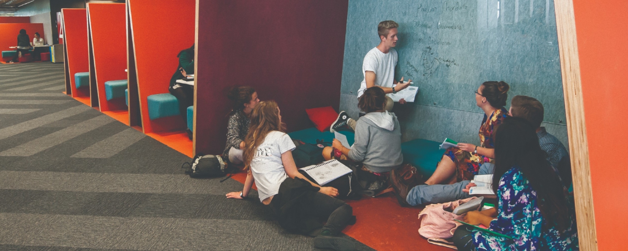 Seven students gather in front of a blue-grey marble wall which one of them is writing on.