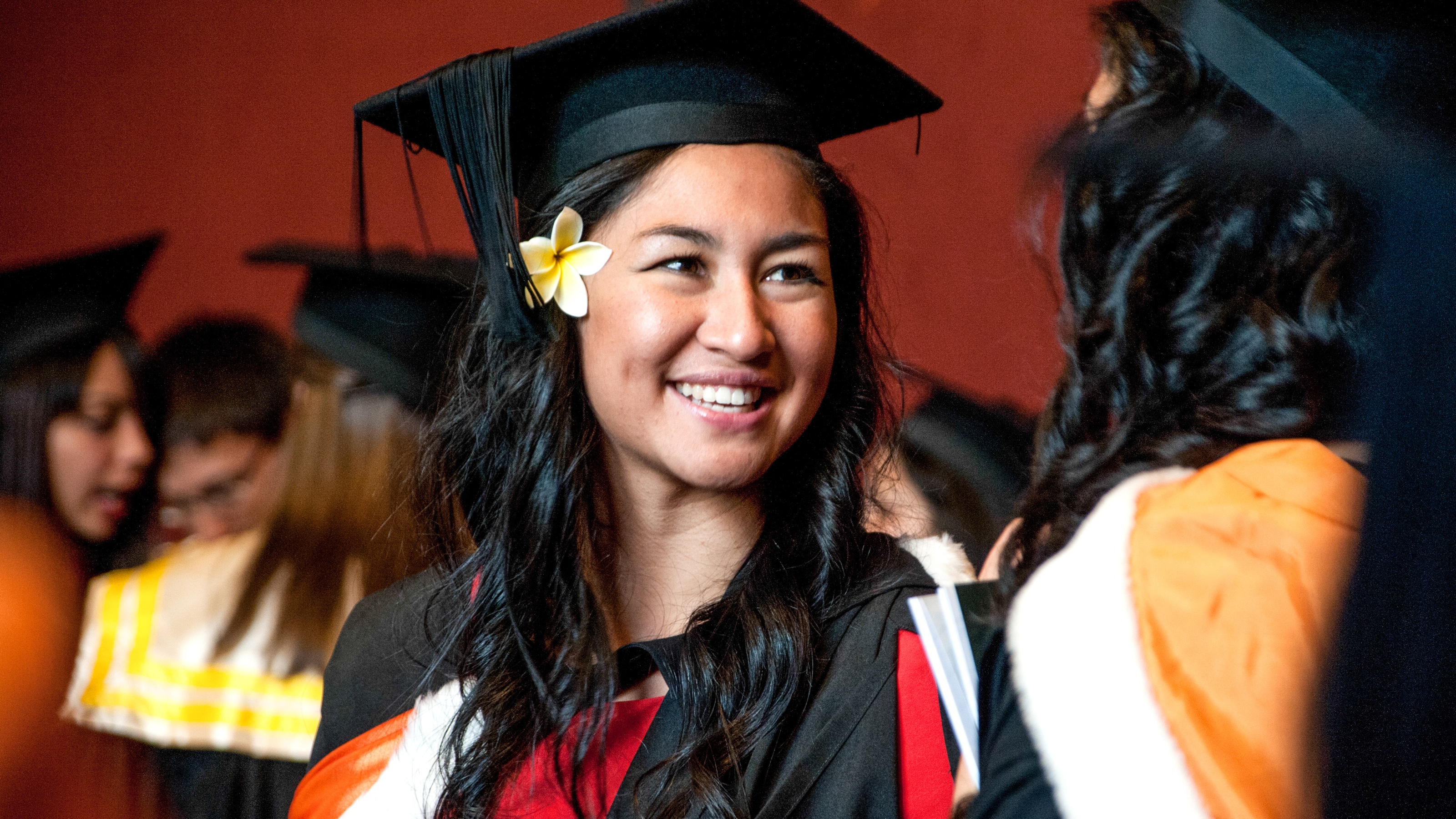 Picture of a young Pasifika woman in graduation robes with a flower in her hair.