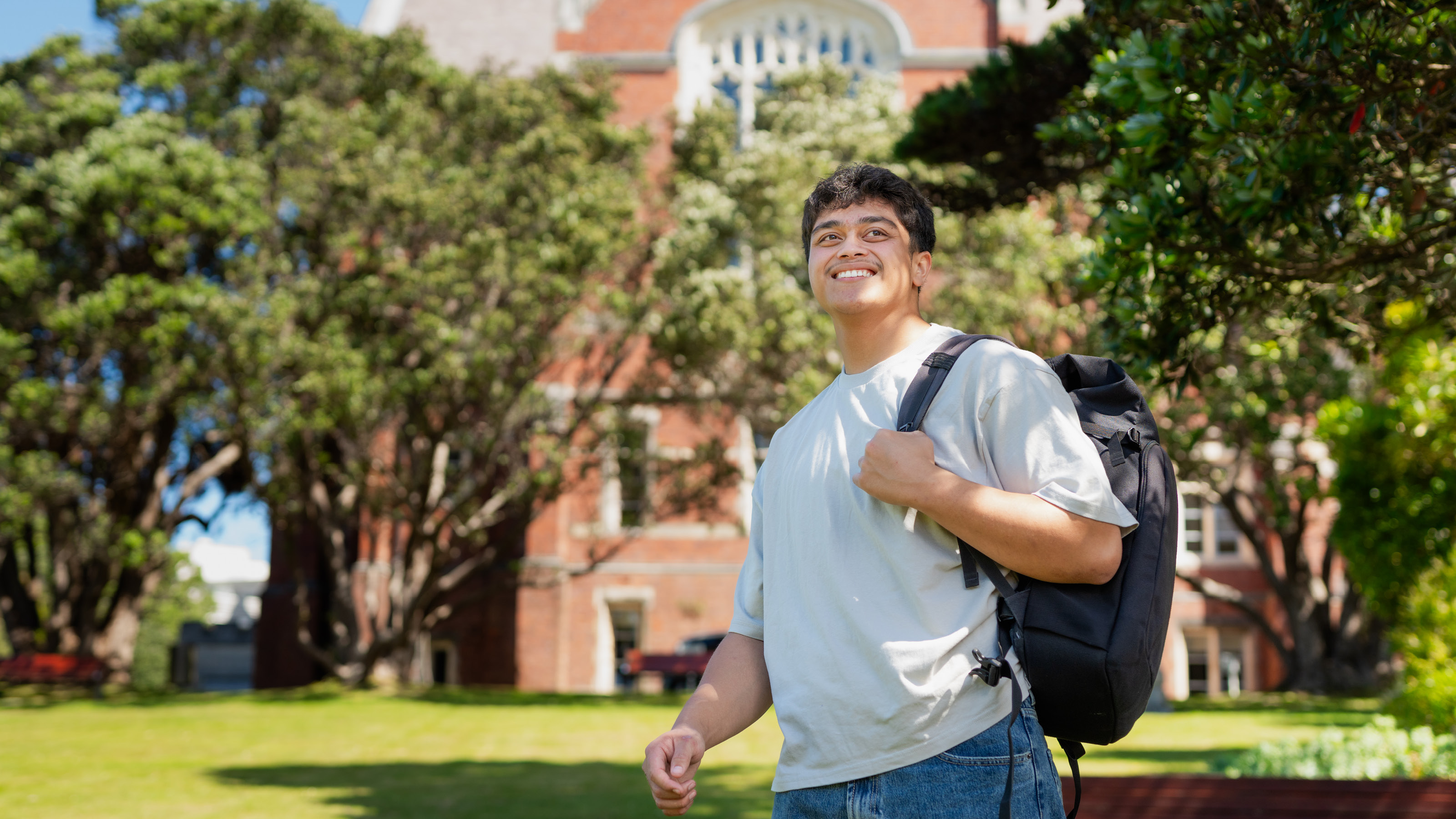 A smiling student stands on the lawn under the trees in front of the Hunter Building
