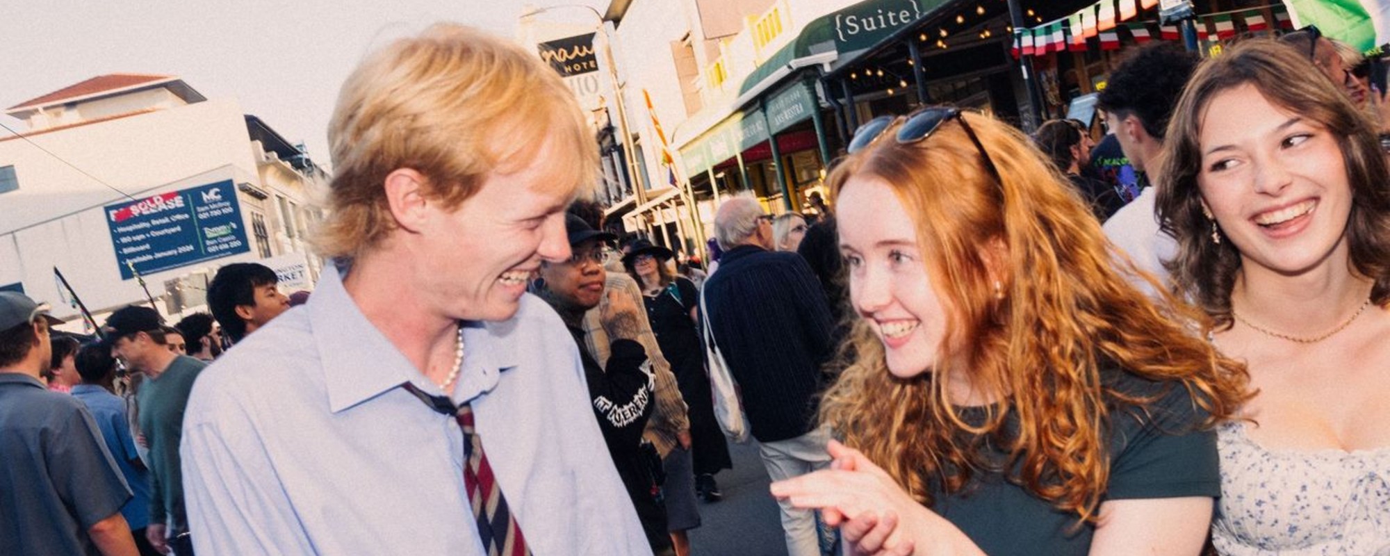 A group of smiling people are gathered outdoors in a busy street setting. In the background, there are shops and buildings, including one with a sign that reads 'Suite' and another displaying flags.